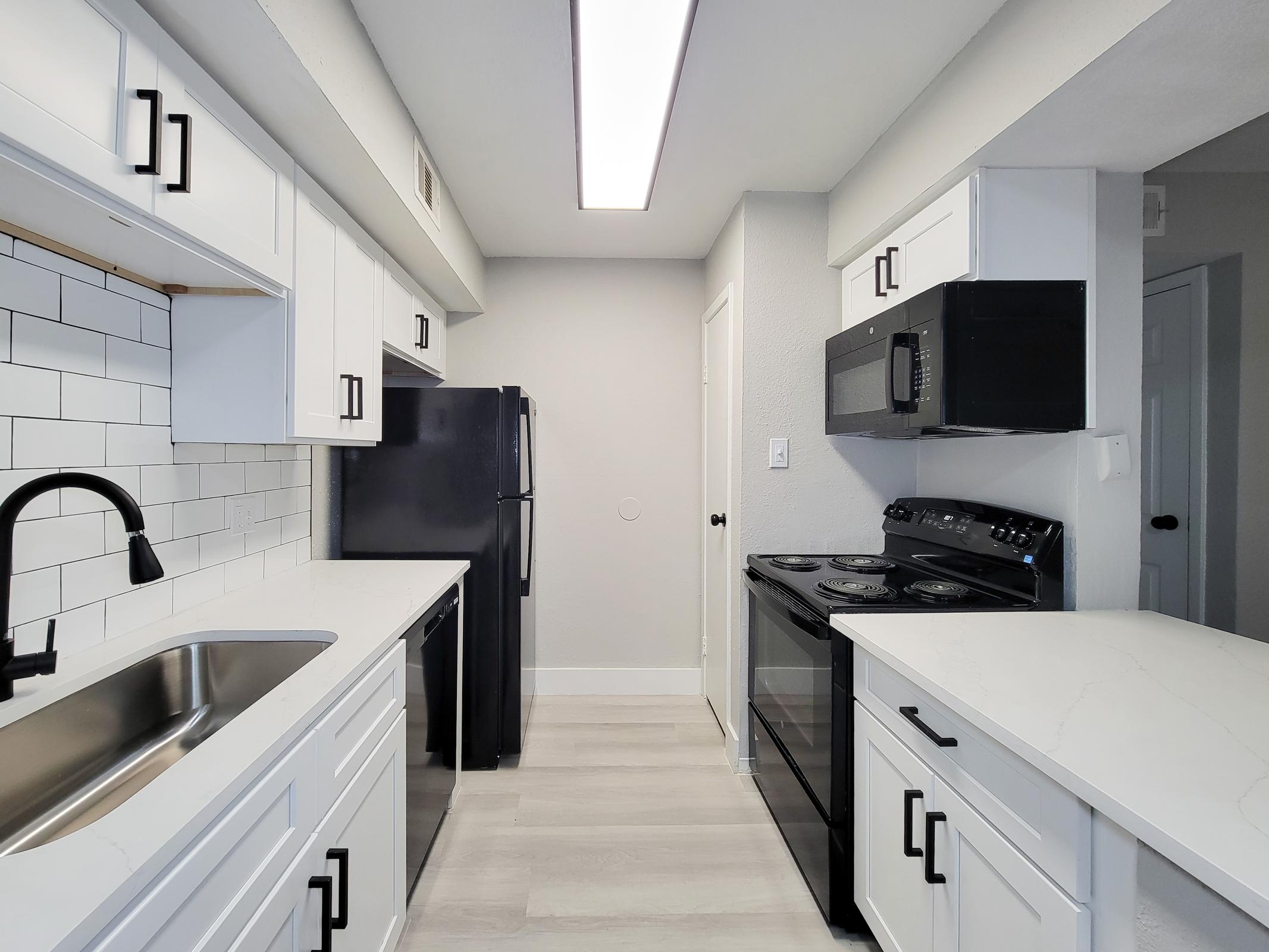 Modern kitchen featuring white cabinets, a black refrigerator, a black microwave, and a black stove. The countertops are light-colored, and there is a stainless steel sink with a black faucet. The backsplash consists of white subway tiles. A bright ceiling light illuminates the space, creating a clean and open atmosphere.