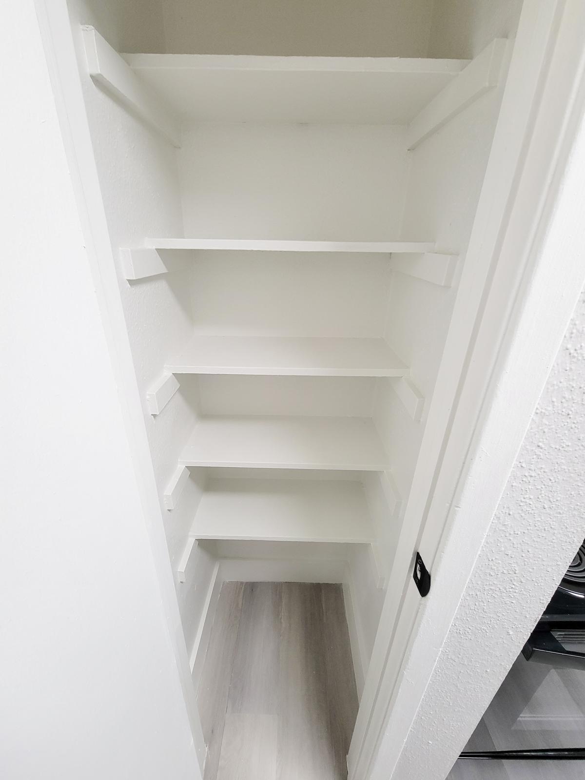 Empty white pantry or storage closet with five shelves and a light-colored floor. The walls and shelves are clean and minimalist, creating a spacious look inside the closet.