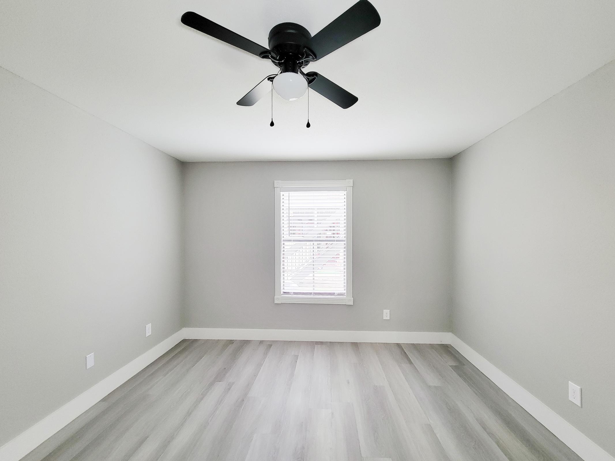 A modern, empty room with light gray walls and a light gray floor. There is a ceiling fan with black blades and a central light fixture. A single window with white blinds allows natural light to enter. The room has a minimalistic and clean design, suitable for various uses.