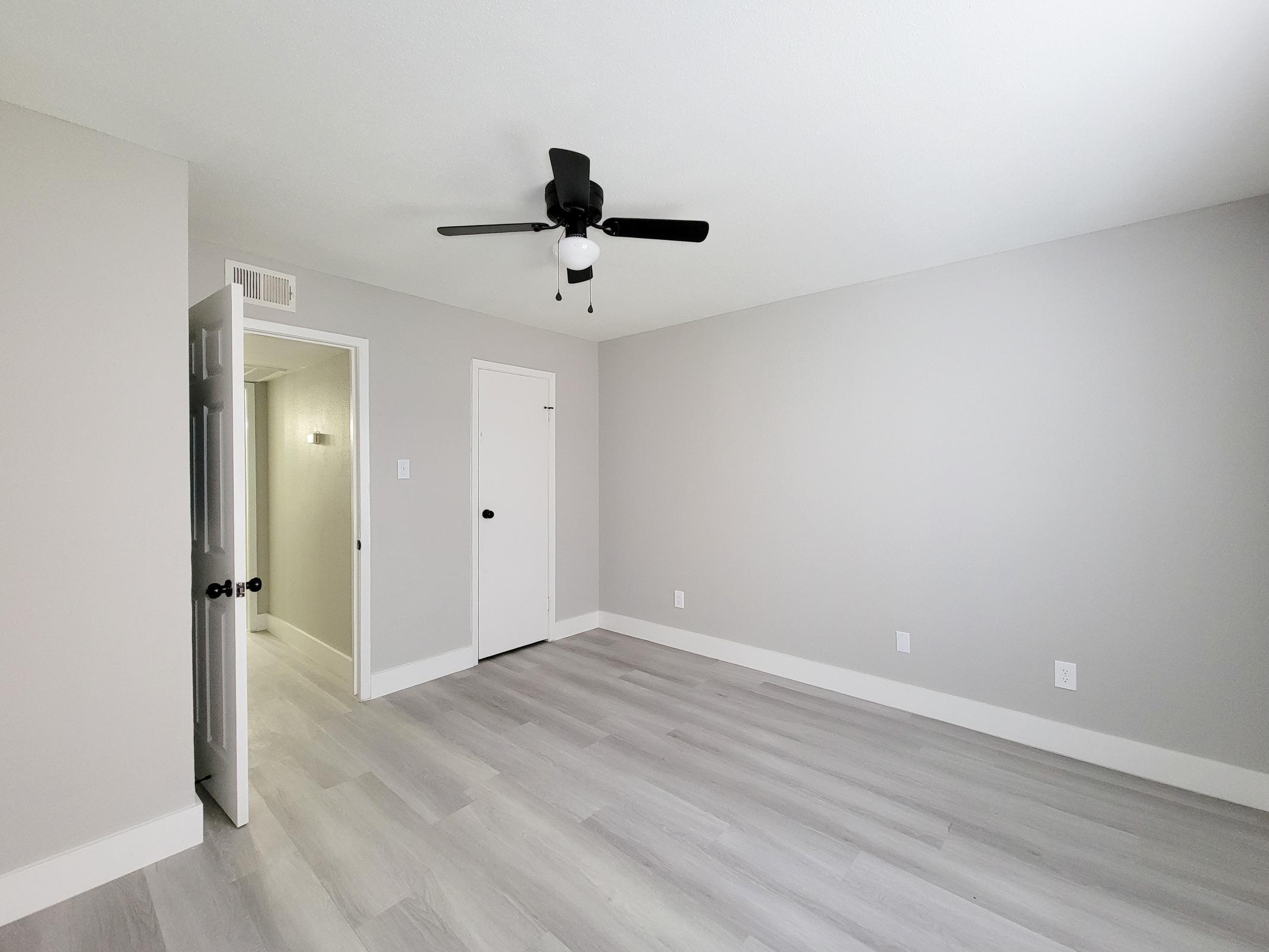 A spacious and minimalistic room featuring light gray walls, a ceiling fan, and light wood flooring. A doorway leads to a hallway, with a closet door visible on the right. The room is bright and uncluttered, creating a calm and inviting atmosphere.