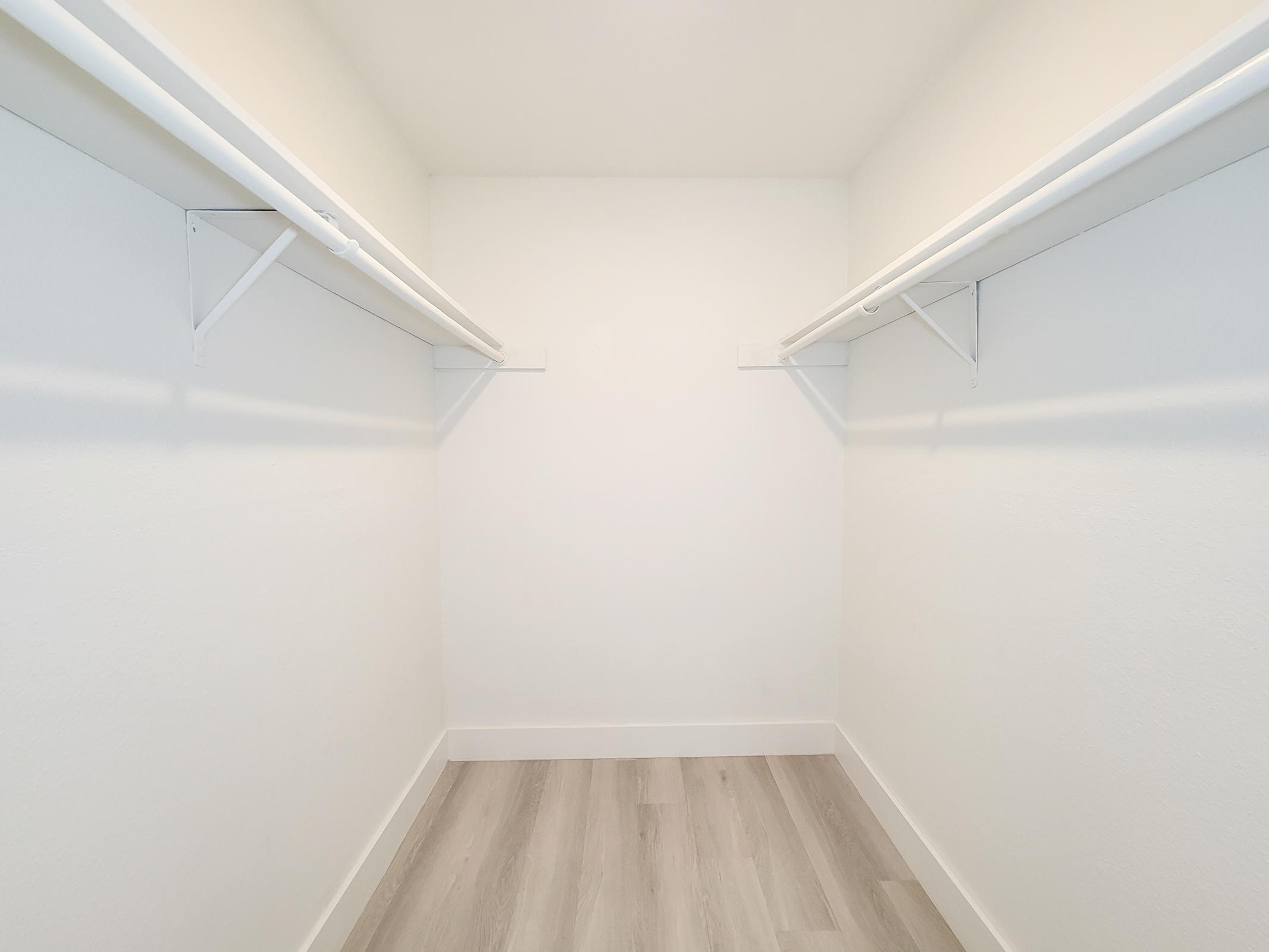 Empty closet with white walls, featuring two white hanging rods on either side. Light-colored wooden flooring adds a modern touch. The space is bright and uncluttered, giving a clean and minimalist look.
