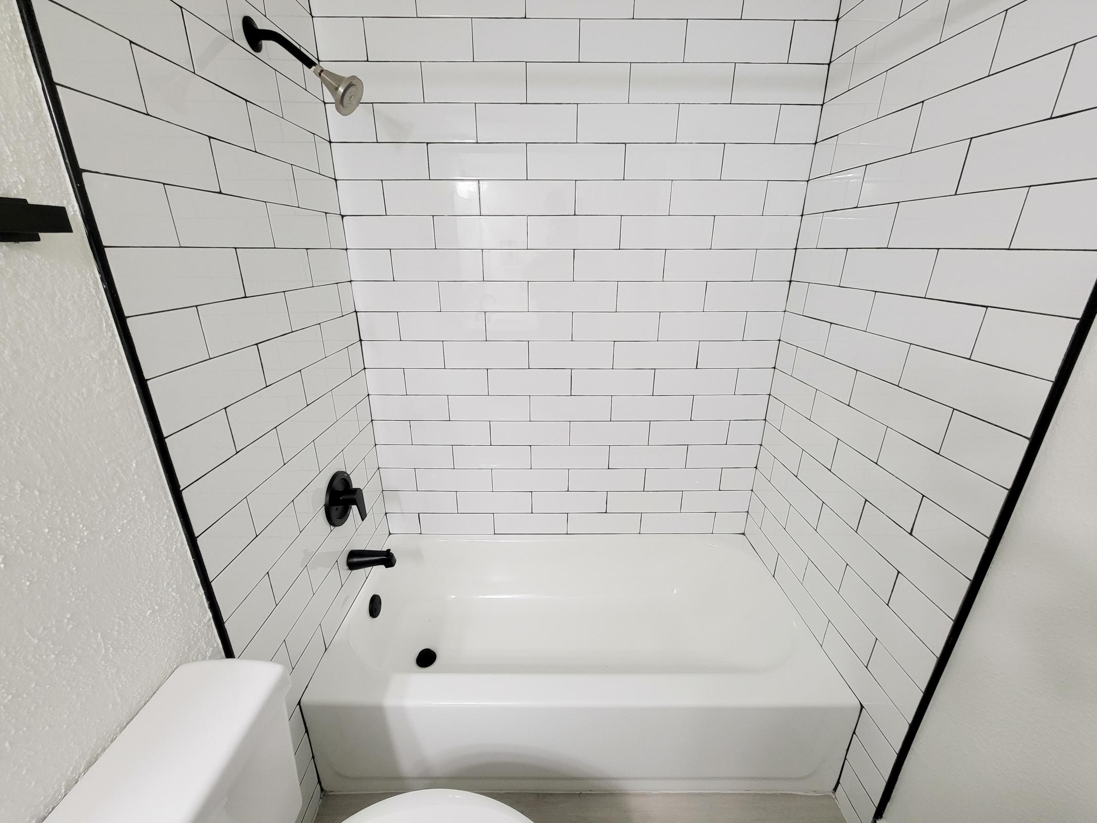 A modern, clean bathroom featuring a white tiled shower with a black accent. The bathtub is sleek and white, positioned against a wall tiled with rectangular white tiles. A black showerhead and faucet contrast with the light-colored tiles, creating a contemporary look.