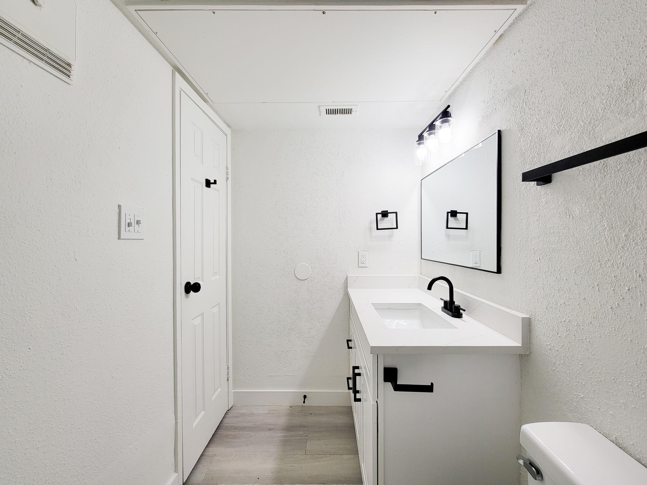 A modern bathroom featuring a white vanity with a sleek countertop, a large mirror above, and stylish black fixtures. The walls are painted white, creating a bright atmosphere, and there is a minimalist shelf and a light fixture with multiple bulbs. A closed bathroom door is visible on the left.