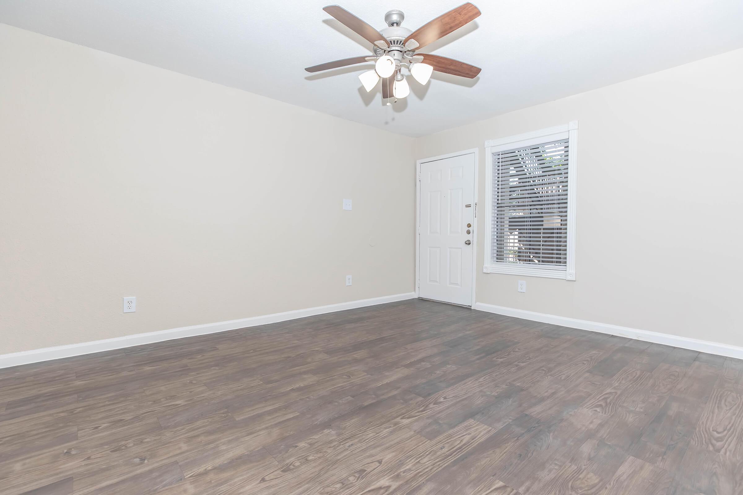 A spacious, empty room with light-colored walls and a ceiling fan. The floor is covered with light brown wood-like laminate. A door is visible on the right leading outside, and a window with white blinds is next to it, allowing natural light to enter the space.