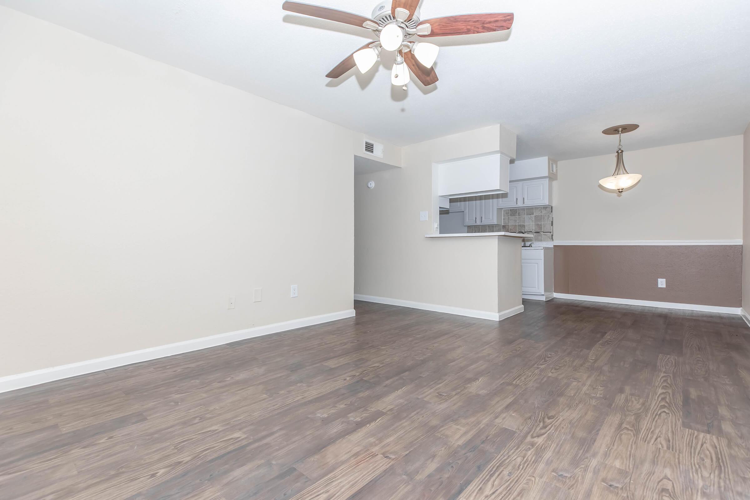 Empty room with light brown laminate flooring, beige walls, and a ceiling fan. A kitchen area is visible in the background with a bar separating it from the living space. There is a pendant light hanging from the ceiling and light fixtures in the room.