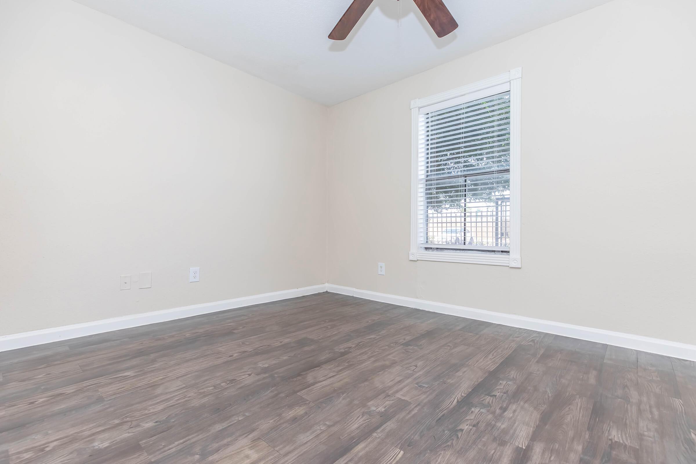 Empty room with light-colored walls and wooden flooring. A window with white blinds is visible, allowing natural light to enter. The ceiling features a brown fan, and the overall space appears clean and spacious.