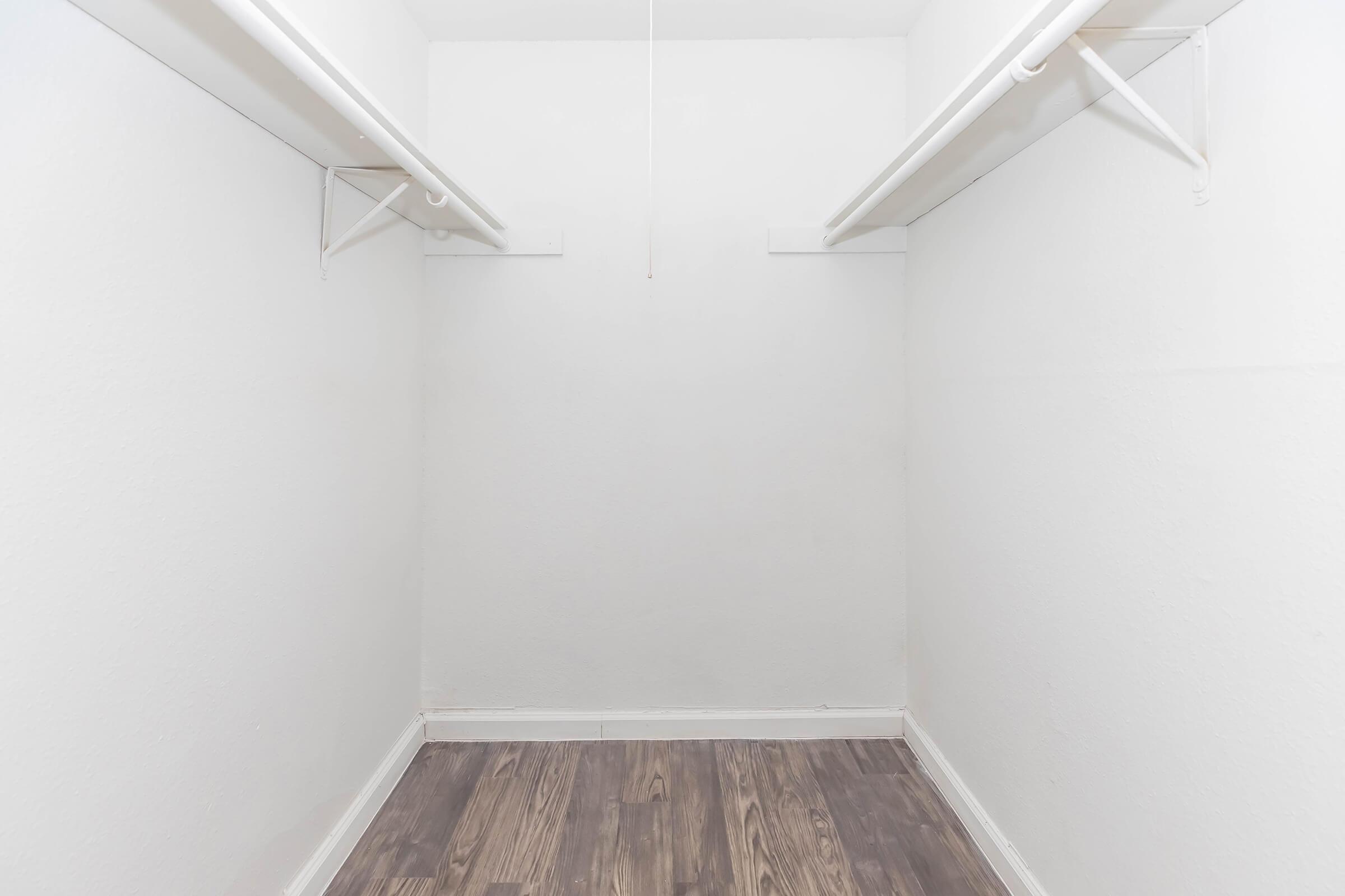 Empty closet with white walls and a wooden floor. There are two horizontal rods along the top for hanging clothes. The space is well-lit and uncluttered, giving a sense of openness.