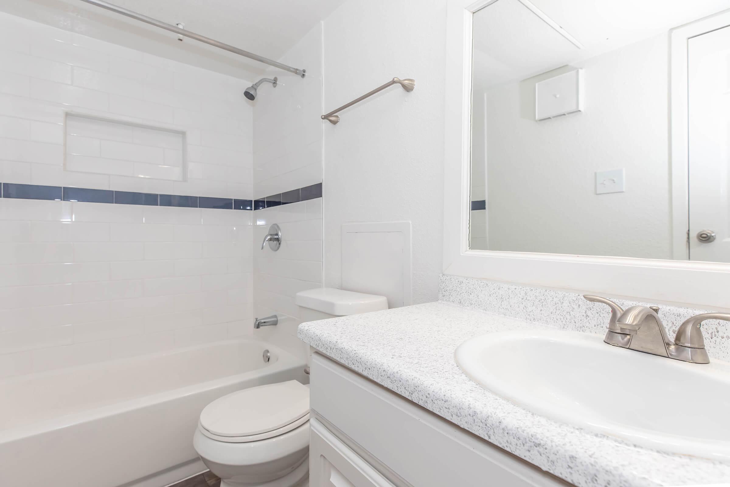 A clean and modern bathroom featuring a white bathtub with a shower, a toilet, and a vanity with a sink. The walls are tiled in white, with a blue accent strip, and there's a large mirror above the sink. Properly lit and well-maintained, the space conveys a fresh and inviting atmosphere.