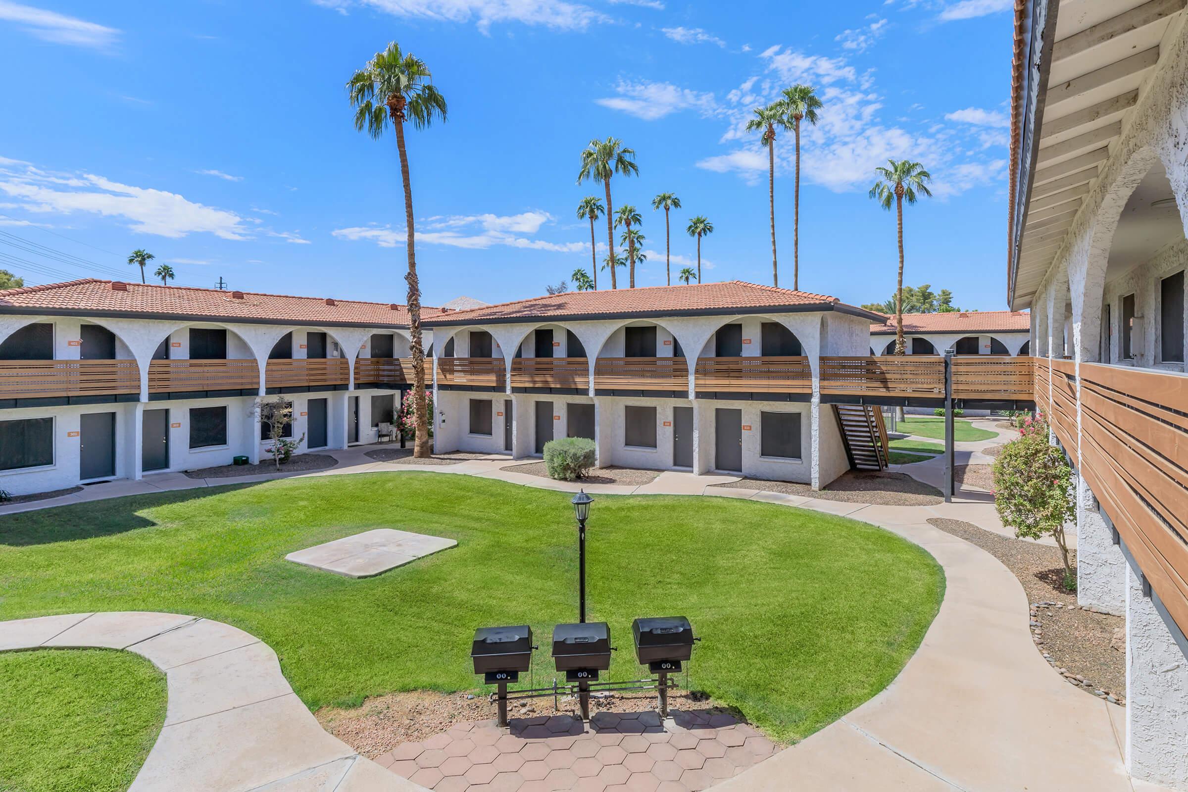 A wide view of a courtyard in an apartment complex featuring lush green grass, palm trees, and several buildings with arched balconies. Grills are positioned on a patio area, and the sky is clear with a few clouds. The setting is well-lit and inviting, showcasing a peaceful residential environment.
