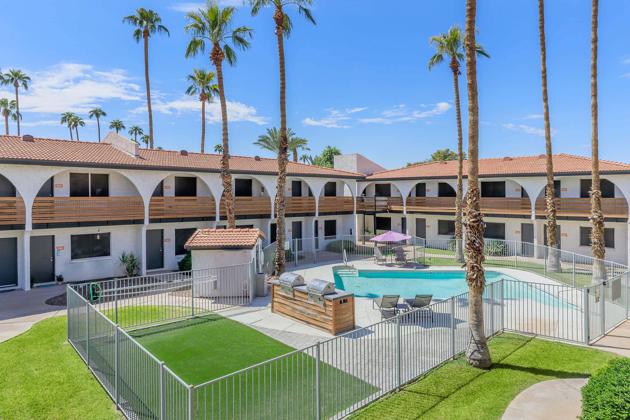 A view of an apartment complex featuring a pool surrounded by palm trees. There are lounge chairs and a shaded area near the pool. The buildings have a modern design with balconies. A well-maintained lawn and a fenced area can also be seen in the foreground, creating a relaxing outdoor atmosphere.