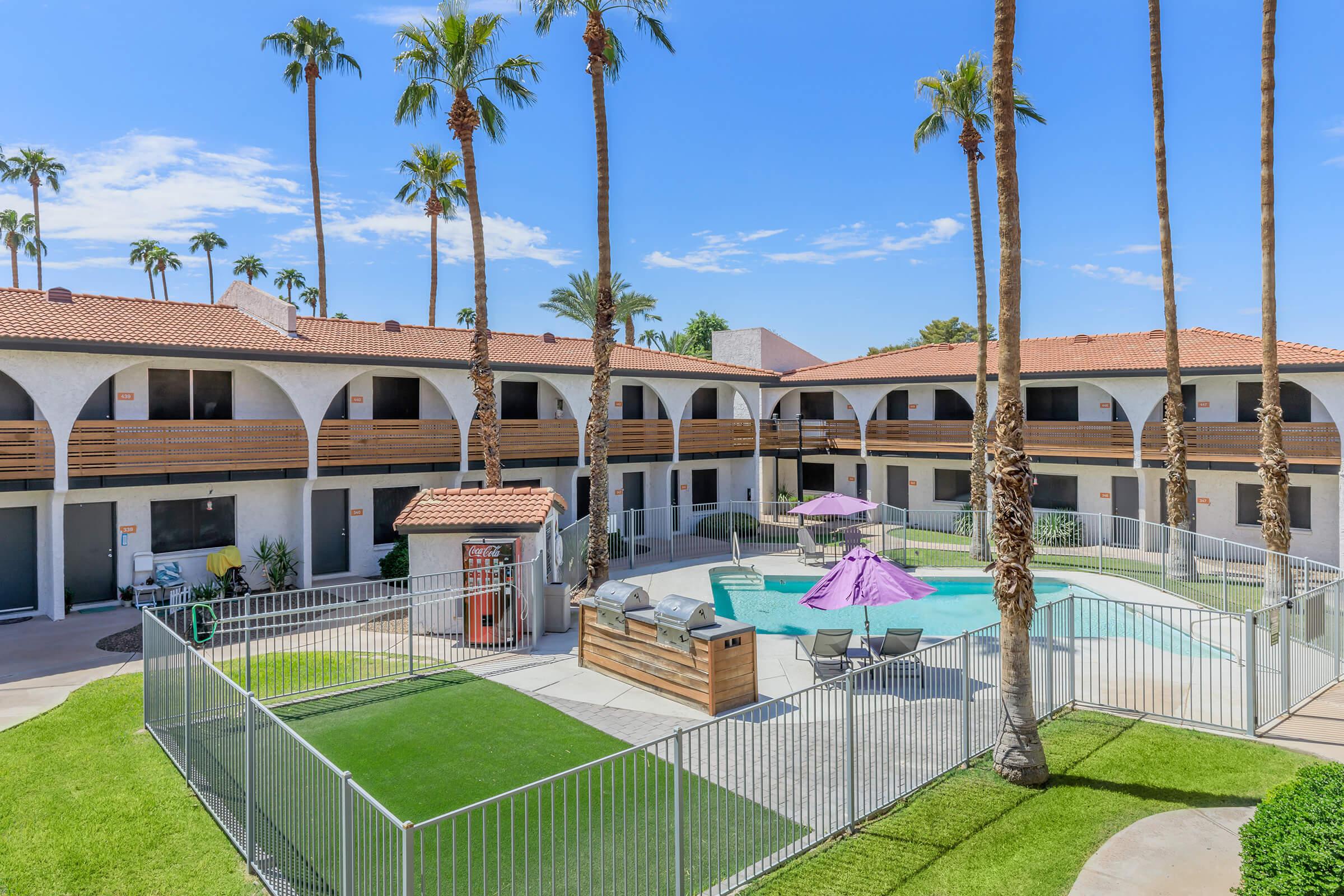 A sunny courtyard of an apartment complex featuring a swimming pool, lounge chairs, and purple umbrellas. Surrounding palm trees add a tropical feel. The buildings have a modern design with arches and balconies, and there is a small fenced area with grass.