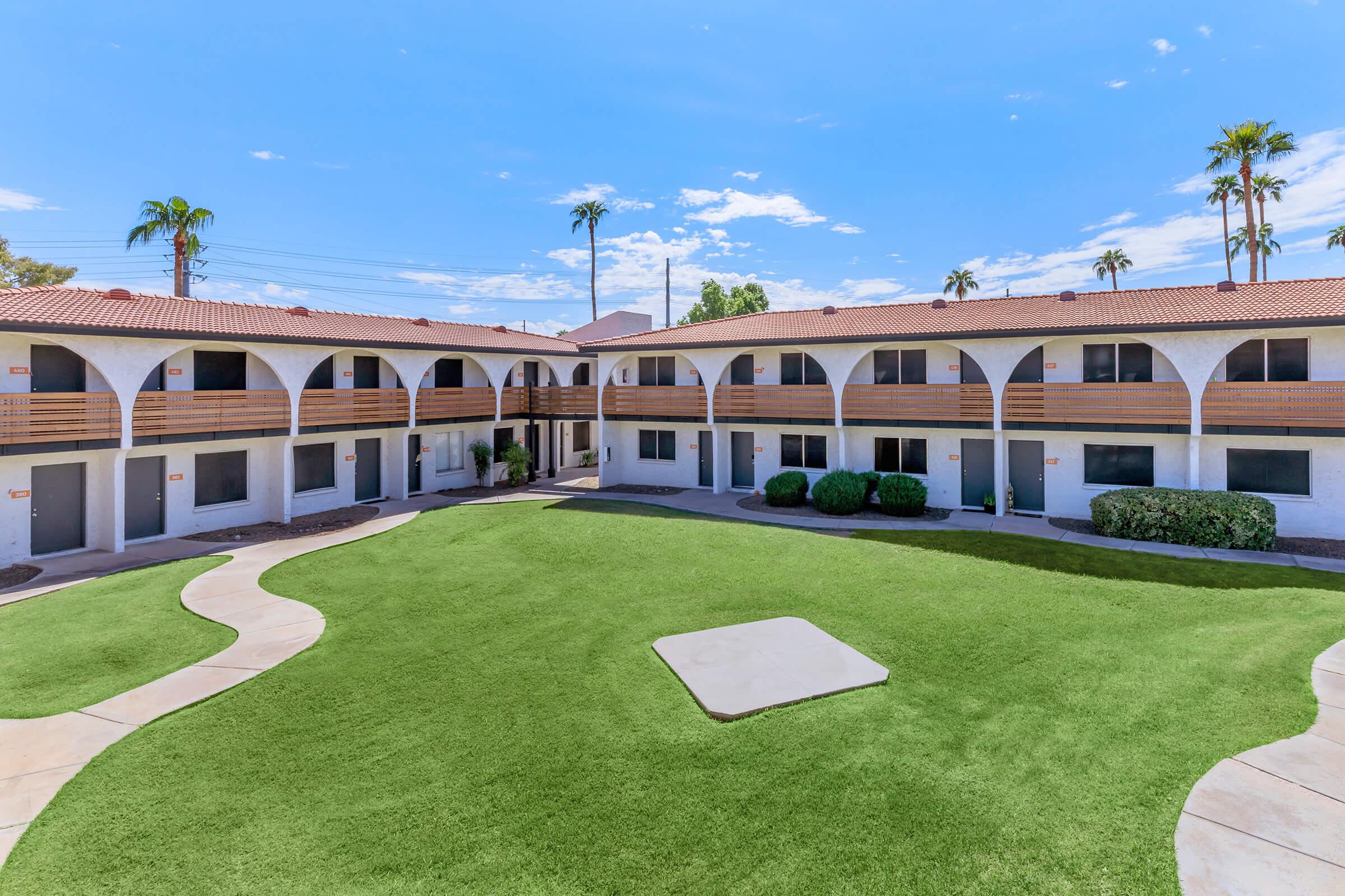 A view of a courtyard in an outdoor hotel or apartment complex. The space features green grass, walkways, and several units with balconies. Palm trees are visible against a blue sky with scattered clouds. The building has an arched architectural style, creating a welcoming atmosphere.