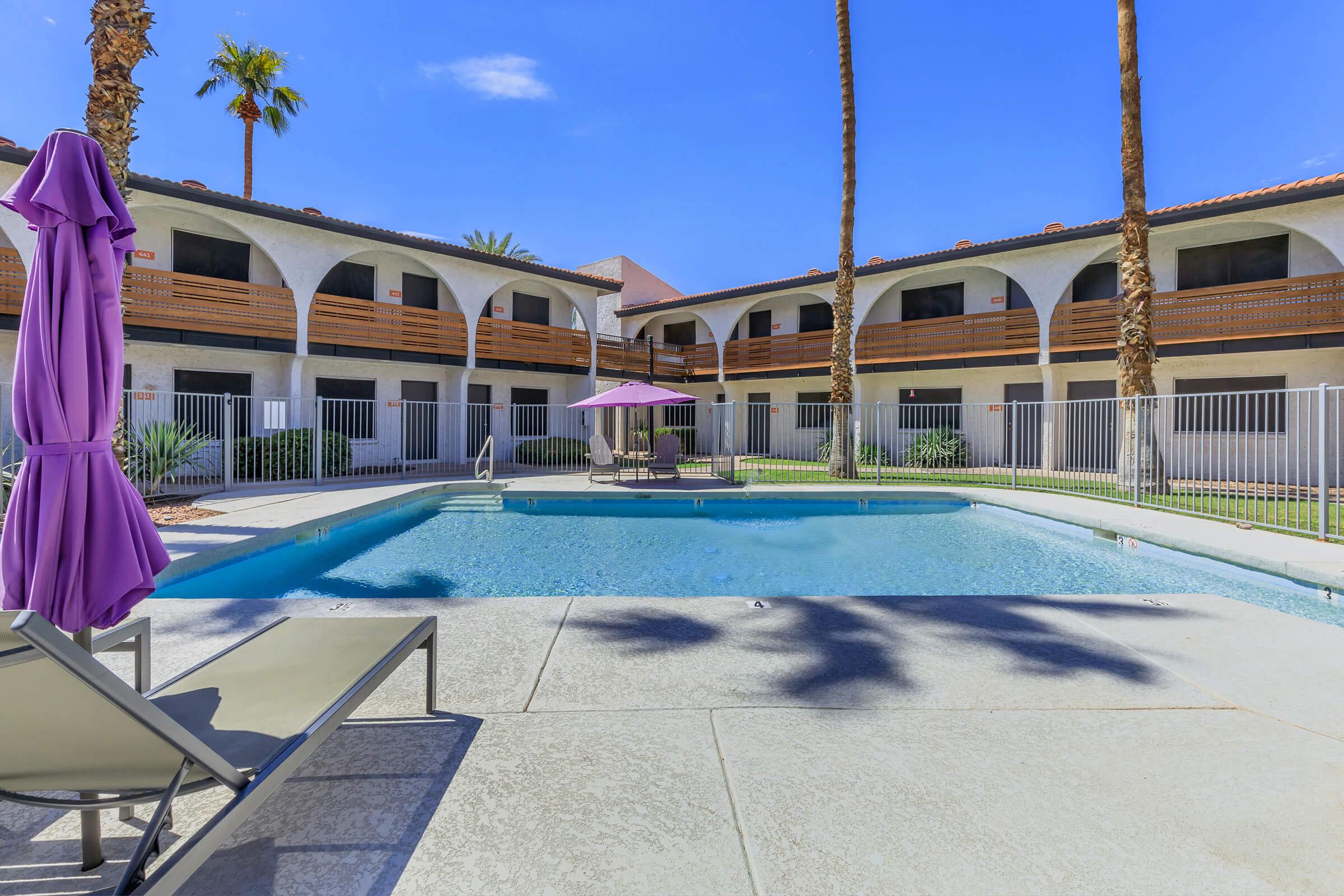 A view of a swimming pool surrounded by lounge chairs and a purple umbrella. Palm trees line the background, with a white building featuring balconies overlooking the pool area. The sky is clear and sunny, creating a relaxing atmosphere.