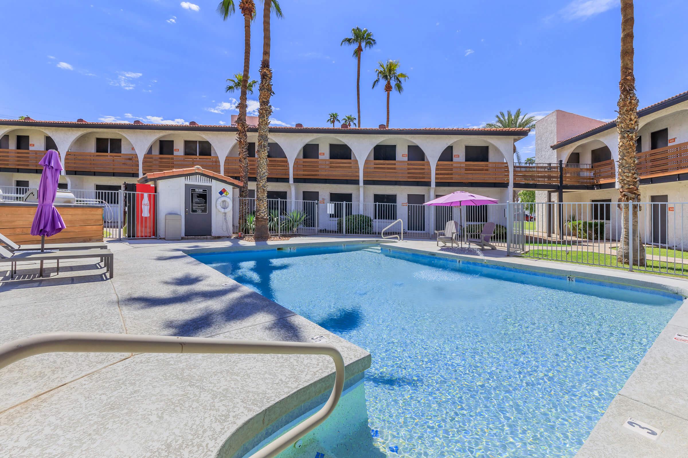 A sunny courtyard featuring a swimming pool surrounded by lounge chairs and palm trees. The area includes two umbrellas, one purple and one pink, and has a two-story building with arched balconies in the background. The scene conveys a relaxed, inviting atmosphere ideal for leisure.