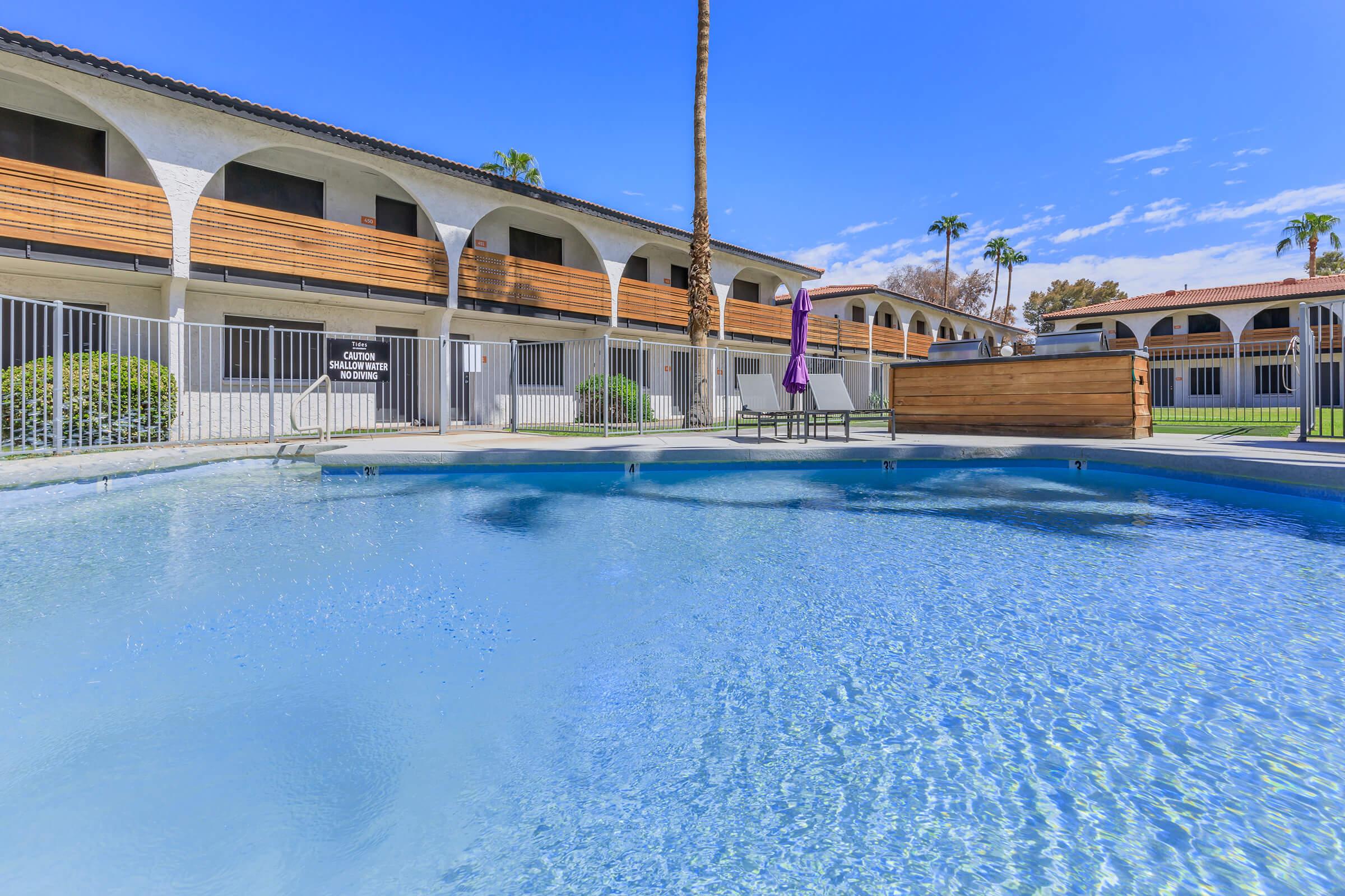 A sparkling blue swimming pool surrounded by a fence, with palm trees in the background and buildings featuring wooden accents. A purple lounge chair sits near the pool, set against a clear blue sky with a few scattered clouds. The area is well-maintained and inviting.