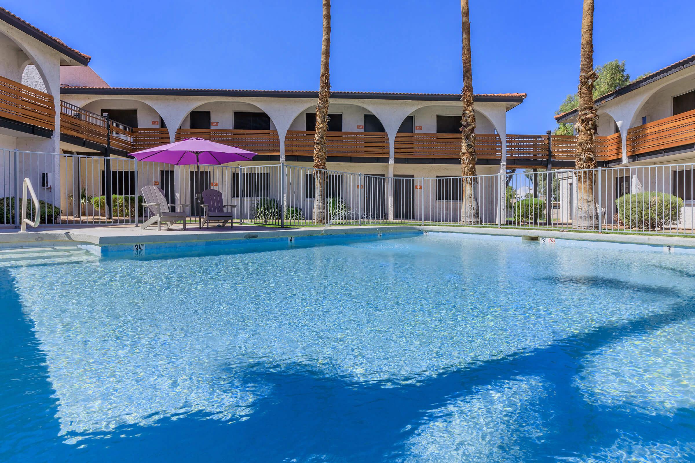 A clear swimming pool surrounded by tall palm trees and an outdoor seating area with a purple umbrella. In the background, there is a two-story building with balconies, and the bright blue sky reflects on the water's surface. The scene conveys a tropical and inviting atmosphere.