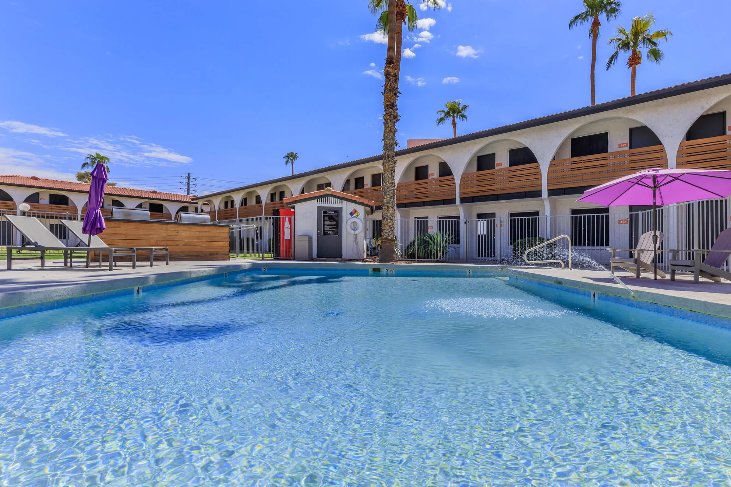 A clear swimming pool surrounded by lounge chairs and umbrellas, with palm trees in the background. The architecture features a two-story building with arched balconies, and bright blue skies above, creating a relaxing outdoor setting.