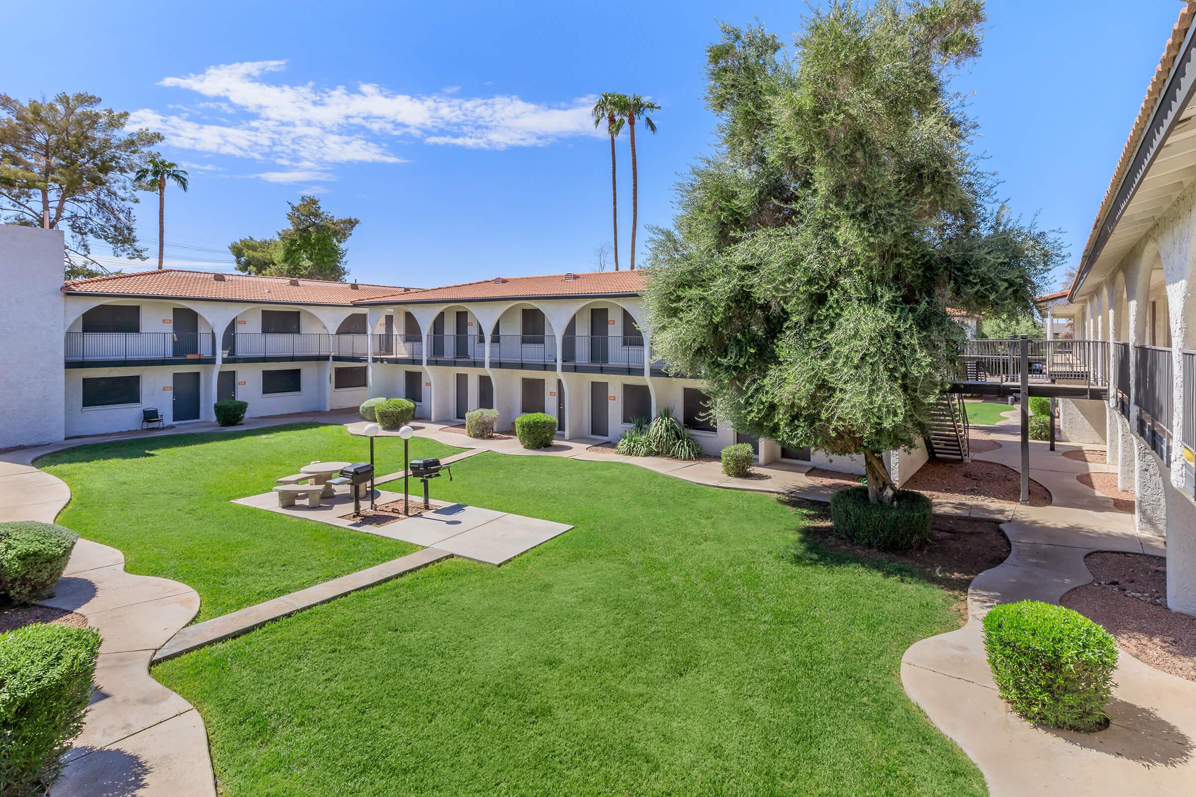 Ground-level view of a landscaped courtyard featuring green grass and shrubs. There are several palm trees and an outdoor grill area with picnic tables. Two buildings with arches surround the courtyard, and the sky is clear with a few clouds.