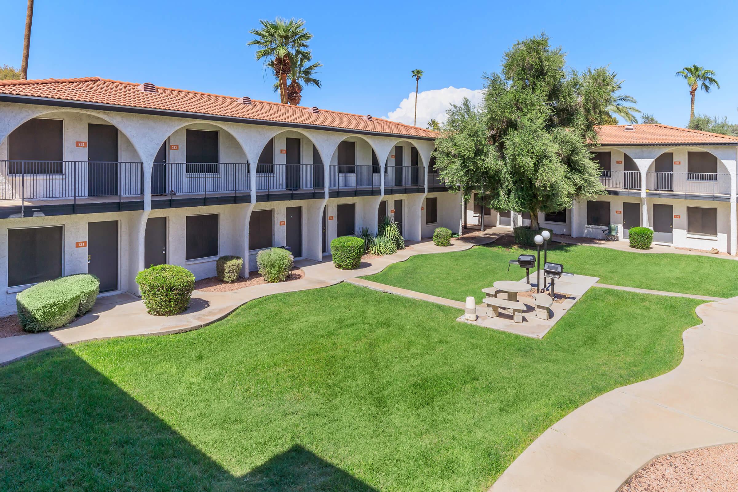 A view of a courtyard in an apartment complex, featuring green grass, shrubs, and palm trees. The building has a tiled roof and multiple arches on the balcony level. There are picnic tables and BBQ grills in the courtyard area, framed by well-maintained landscaping under a clear blue sky.