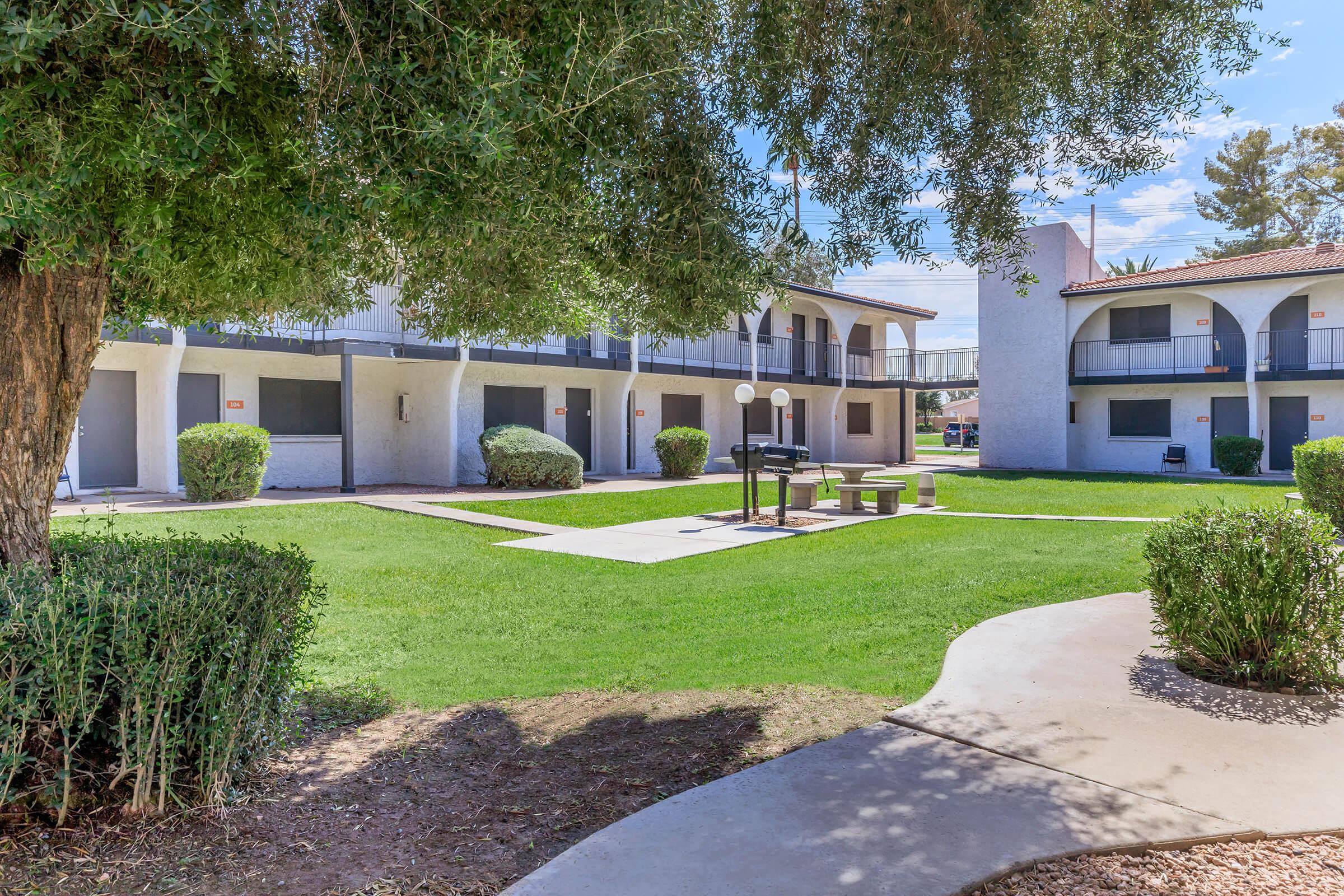 A well-maintained outdoor area featuring a green lawn, bushes, and a paved pathway. There are two buildings with balconies in the background and a communal seating area with a grill. The setting is bright and inviting, with trees providing shade.
