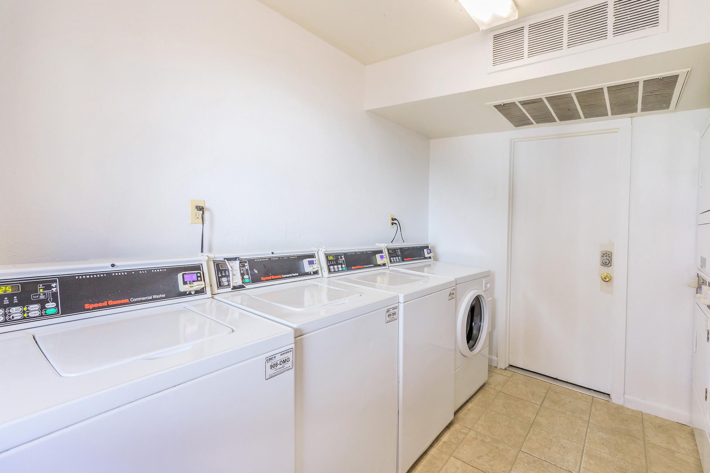 A clean and tidy laundry room featuring several white washing machines and a dryer lined up against a wall. The floor is tiled, and there is a door that leads outside. The walls are painted white, contributing to a bright and fresh atmosphere in the space.