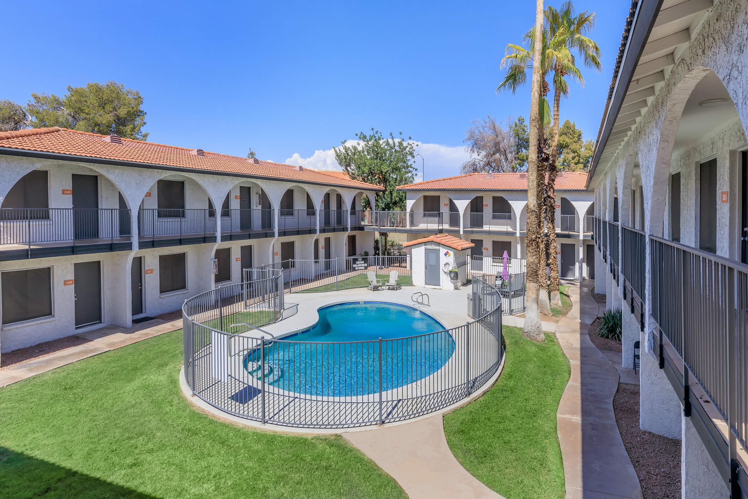 A view of a hotel courtyard featuring a swimming pool surrounded by a fenced area. The pool area is bordered by green grass and palm trees, with two-story buildings and arched walkways in the background under a clear blue sky.