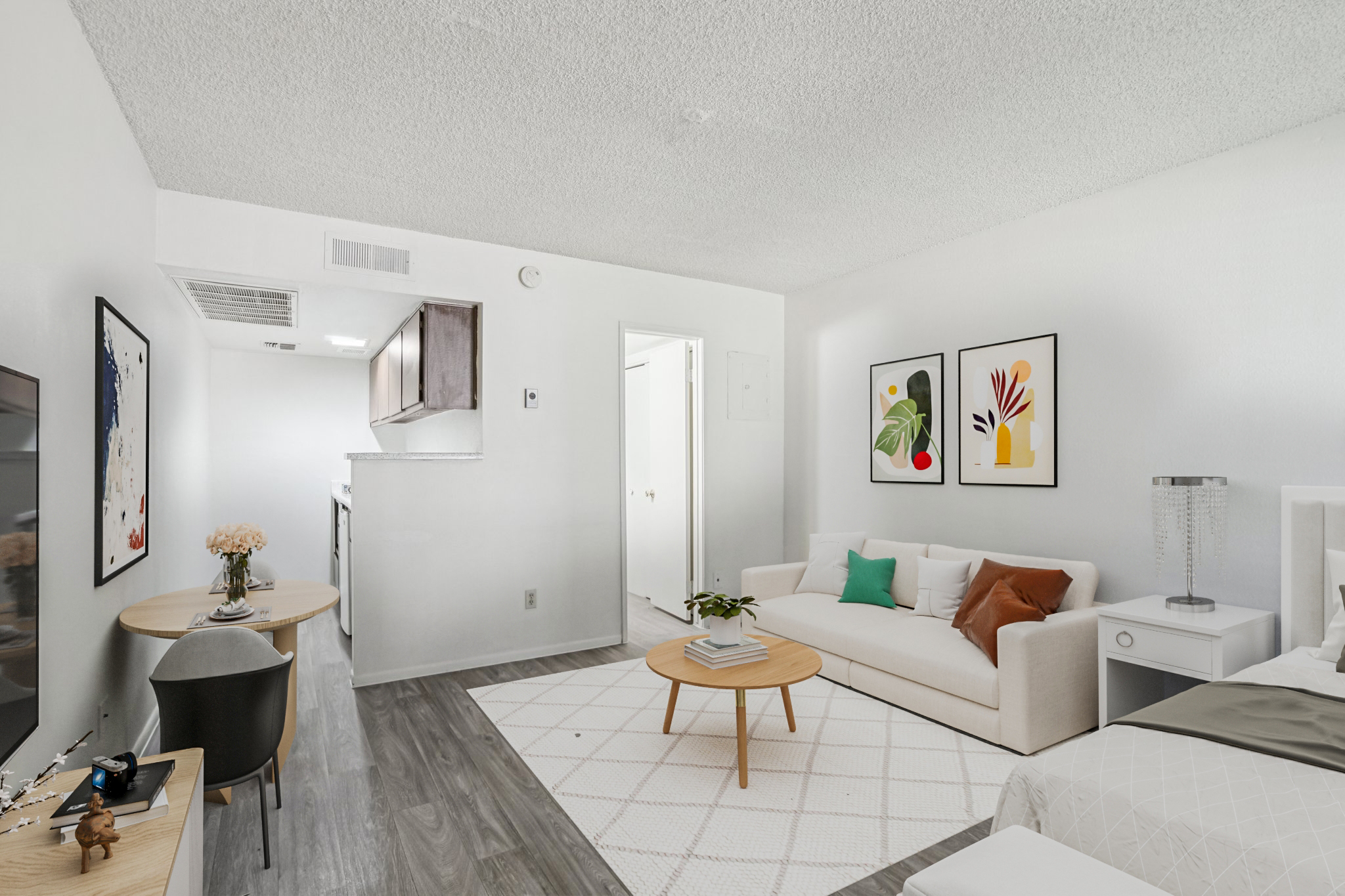 A bright and modern living space featuring a white couch adorned with colorful pillows, a round coffee table, and a cozy area rug. A small dining table is visible in the background, along with a kitchenette. Two framed art pieces hang on the wall, adding a touch of decor to the minimalist setting.
