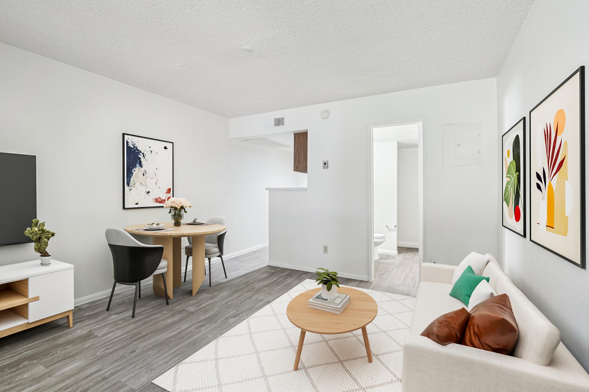 A bright, modern living space featuring a light-colored sofa with brown cushions, a round coffee table, and a dining area with a circular table and two chairs. Decorative wall art adorns the walls, and a small plant adds a touch of greenery. The room has a clean, minimalist design with neutral tones.