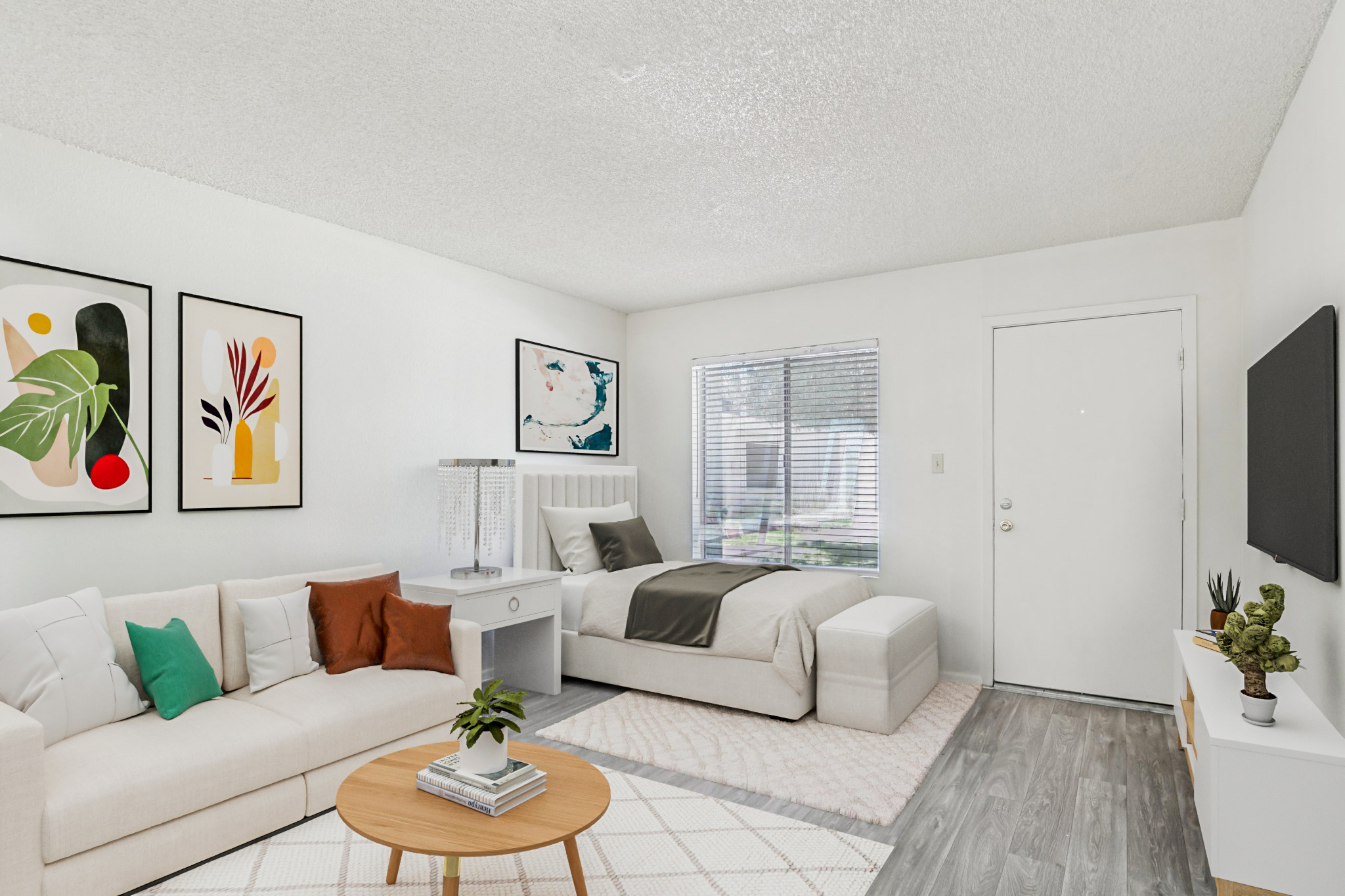 A modern, cozy living space featuring a light-colored sectional sofa with colorful throw pillows, a round wooden coffee table, and a bed with neutral bedding. The walls are decorated with framed artwork, and there's a window letting in natural light. A television is mounted on the opposing wall, and a welcoming entrance is visible.
