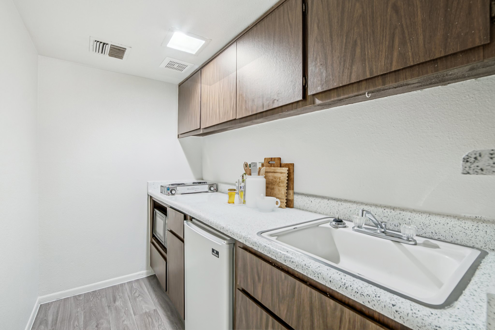 A modern kitchen interior featuring wooden cabinets, a countertop with a sink, a small refrigerator, and a stovetop. The space is well-lit with ceiling lights and has a clean, minimalist design, showcasing a neutral color palette and a few kitchen essentials on the counter.
