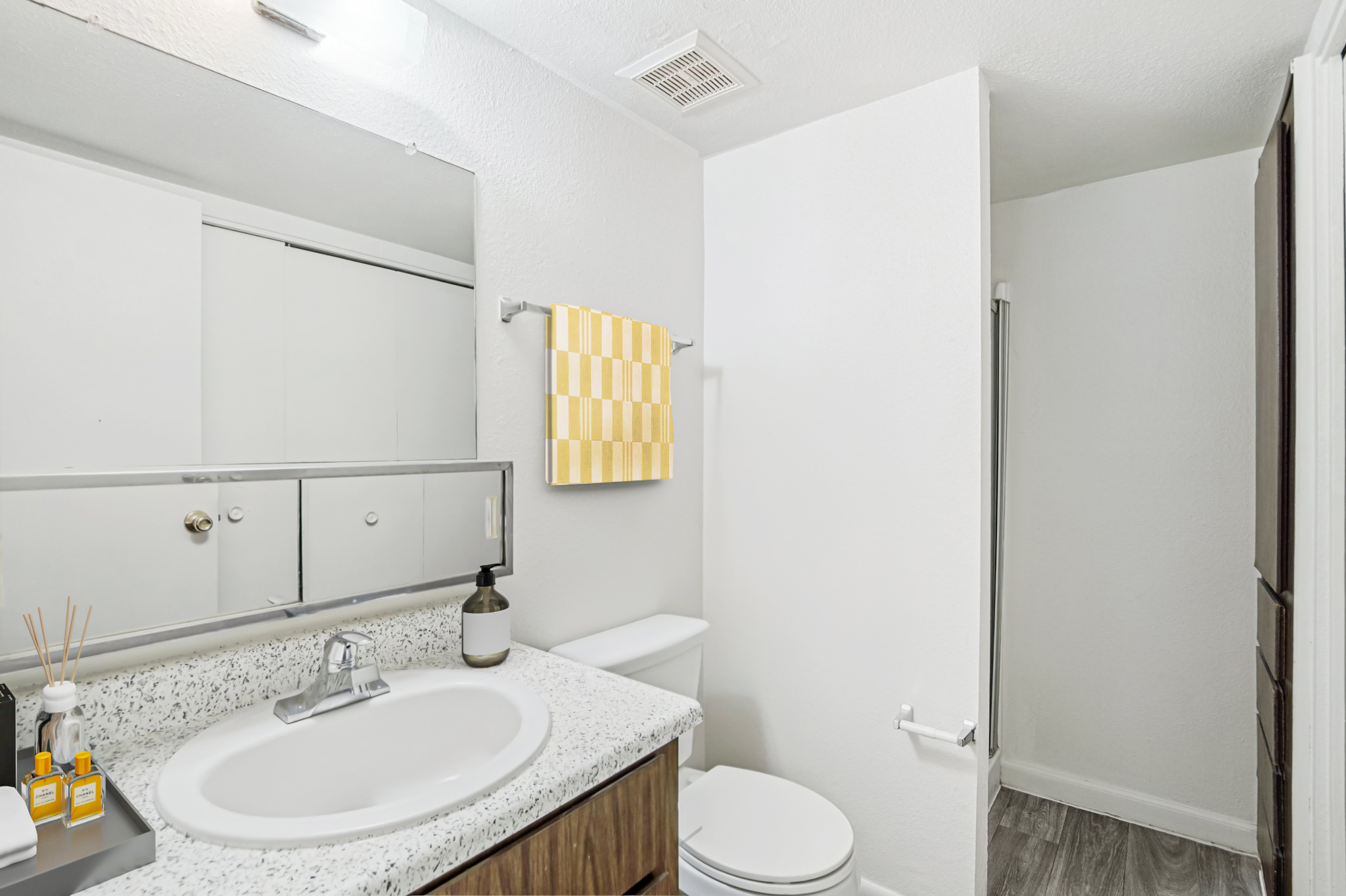 A modern bathroom featuring a light-colored granite countertop, a round sink, a toilet, and a mirror with a simple design. The walls are painted white, and there is a yellow and white checkered towel hanging. Decorative items, including a small bottle and reed diffuser, are placed on the countertop. A door leads to a separate area.