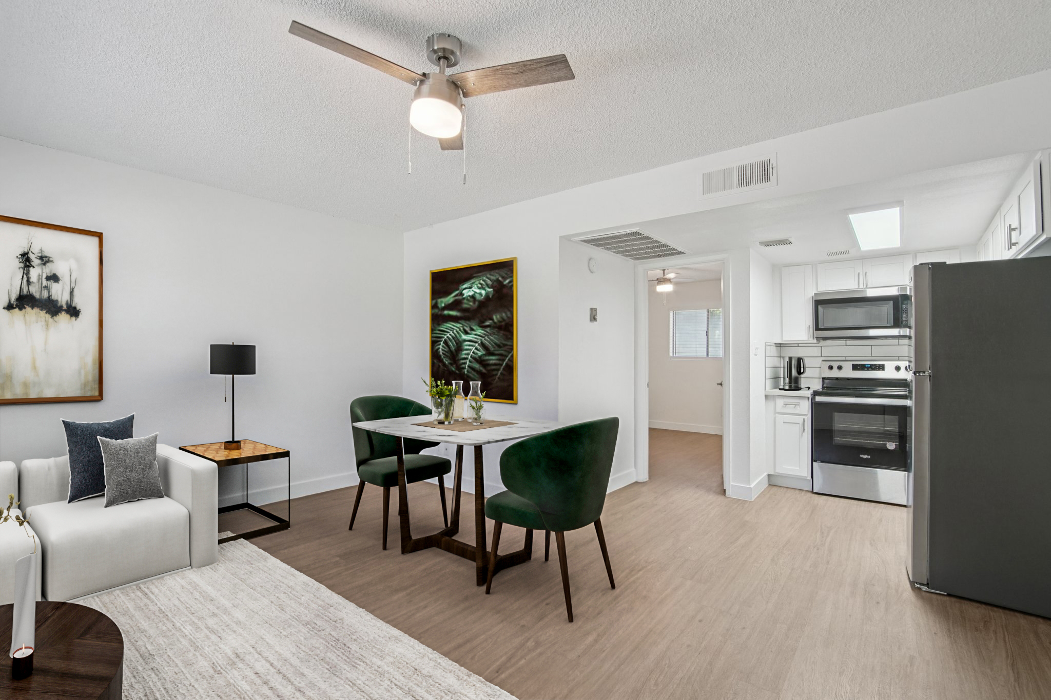 A modern and airy living space featuring a white sofa, a small dining table with green chairs, and a ceiling fan. The decor includes a framed artwork on the wall, with a view into a kitchen area equipped with stainless steel appliances, all set against light-colored walls and flooring.