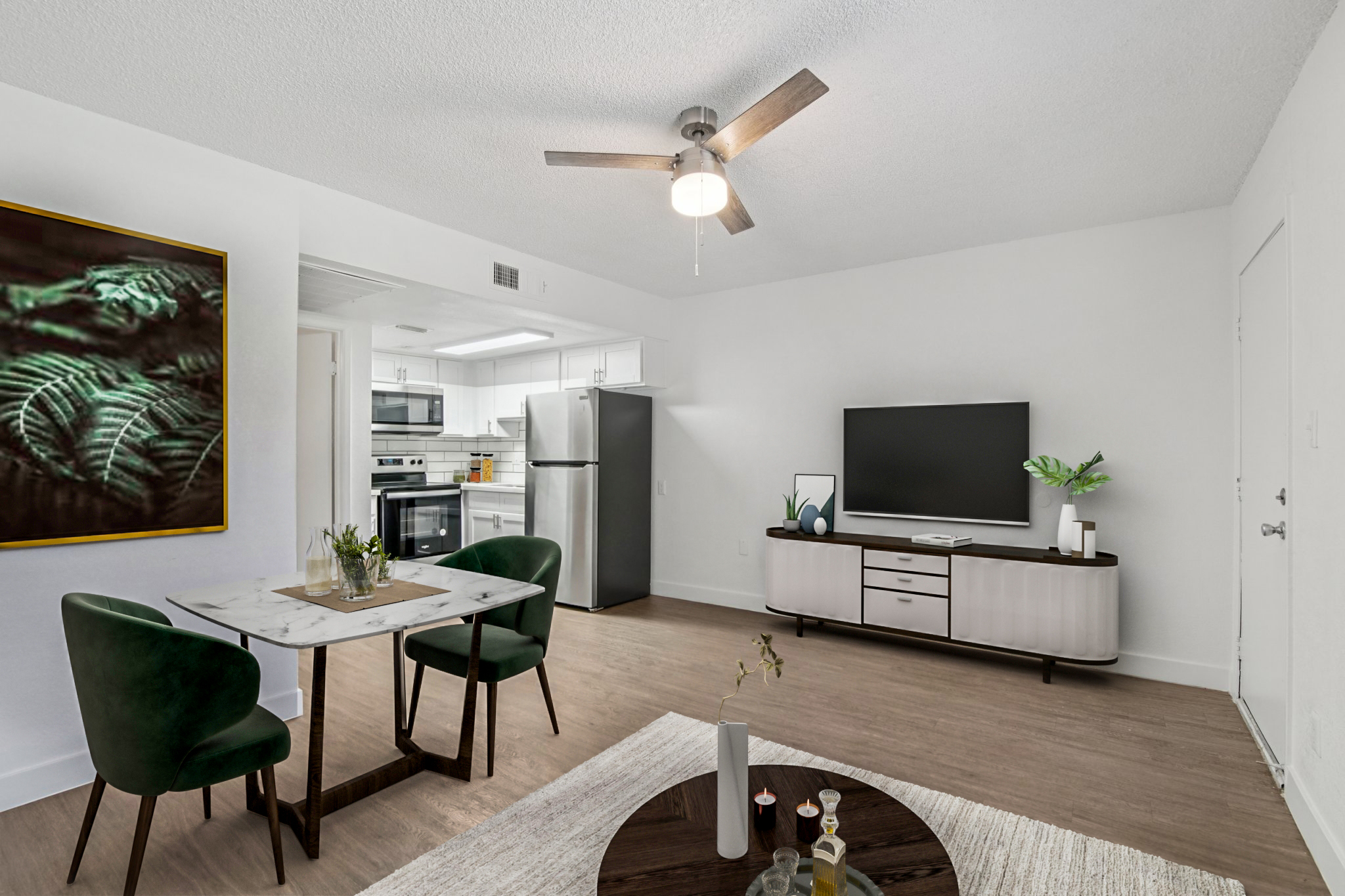 Modern living room and dining area featuring a small round table with green velvet chairs, a large flat-screen TV on a light wood media console, and a ceiling fan. A kitchen with stainless steel appliances is visible in the background, along with decorative elements like a plant and a rug.