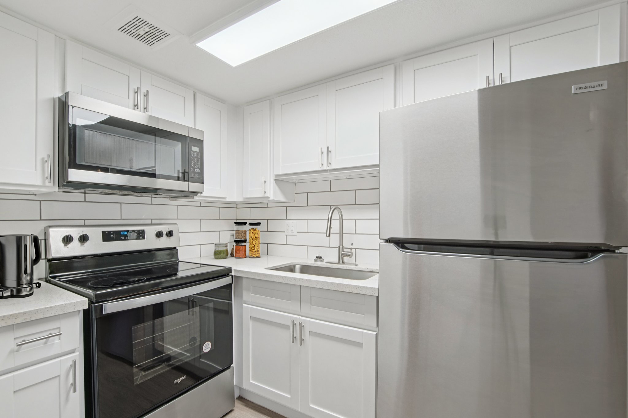 Modern kitchen featuring stainless steel appliances, including a microwave, oven, and refrigerator. White cabinetry complements a light countertop and a tile backsplash. A stainless steel sink is positioned between the stove and fridge, with a coffee maker and jars on the countertop, creating a clean and functional space.