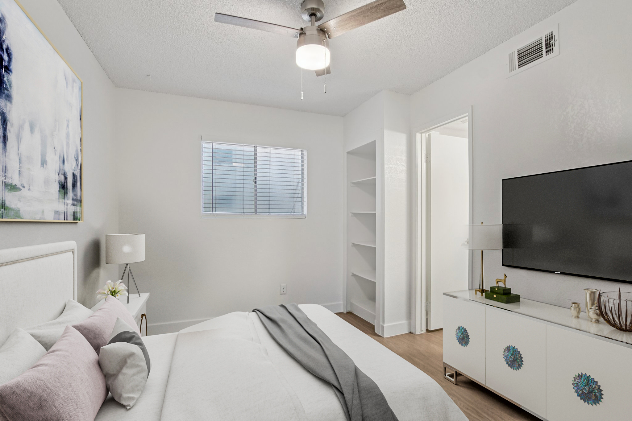 A modern bedroom featuring a white bed with decorative pillows, a ceiling fan, a window with blinds, and a wall-mounted TV. There is a light-colored dresser with a decorative motif and a small shelf unit. Soft, neutral tones create a calm and inviting atmosphere.