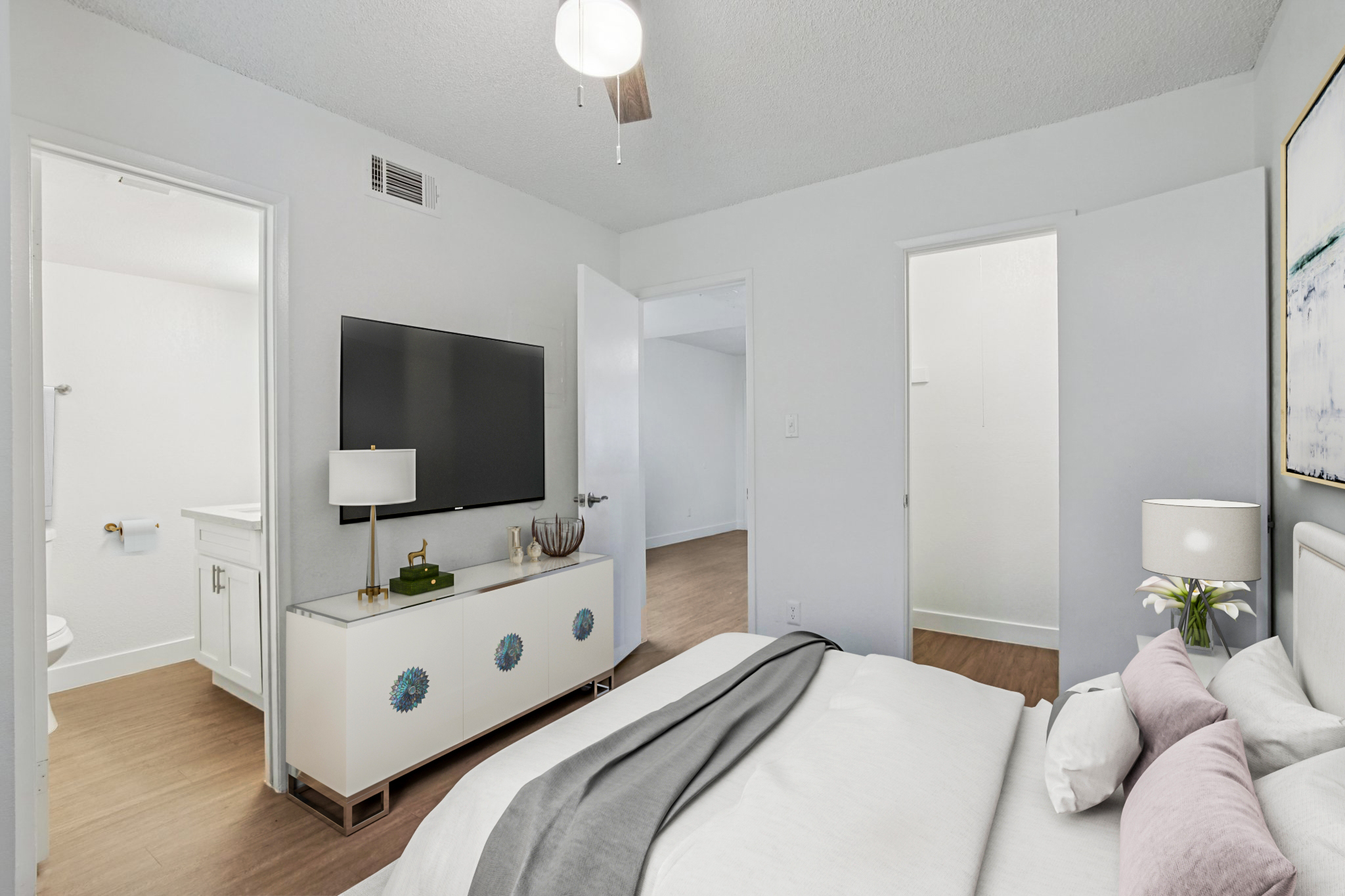 A modern bedroom featuring a bed with white linens and decorative pillows, a flat-screen TV on a light-colored dresser, a ceiling fan, and two open doors leading to a bathroom and another room. The walls are painted white, and the flooring is wooden, creating a bright and airy atmosphere.