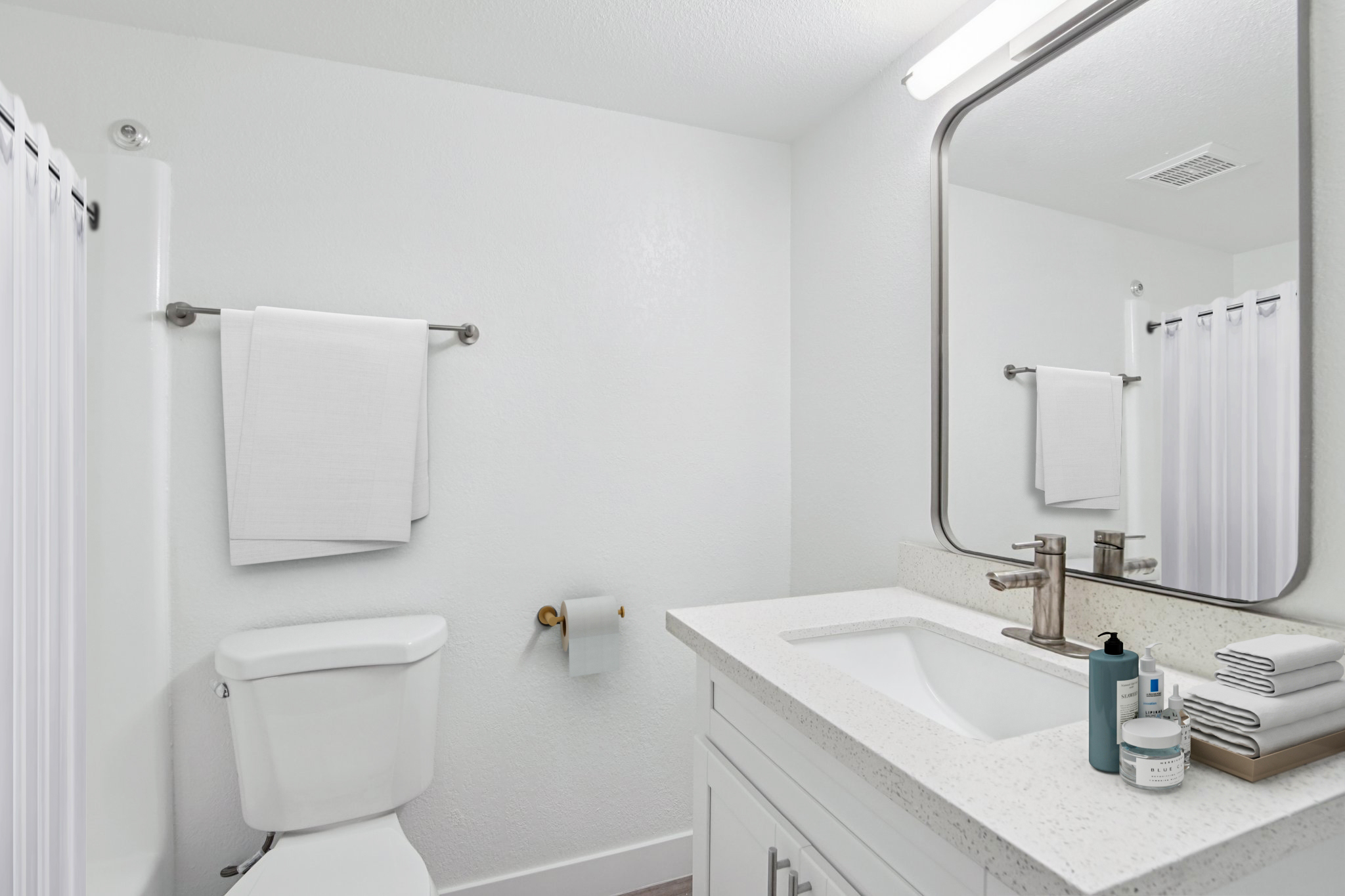 Bright and modern bathroom featuring a white toilet, a large mirror above a sleek countertop with a sink, and neatly arranged towels. The space is decorated with muted colors, has a curtain for the shower, and includes toiletries on the countertop, creating a clean and inviting atmosphere.