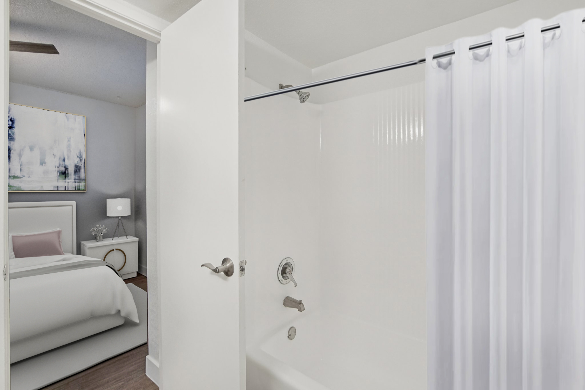 A view of a bathroom showing a shower/tub combo with a white curtain, adjacent to a bedroom visible through an open door. The bedroom features a bed with light-colored linens and a nightstand with a lamp, set in a softly lit, modern environment.