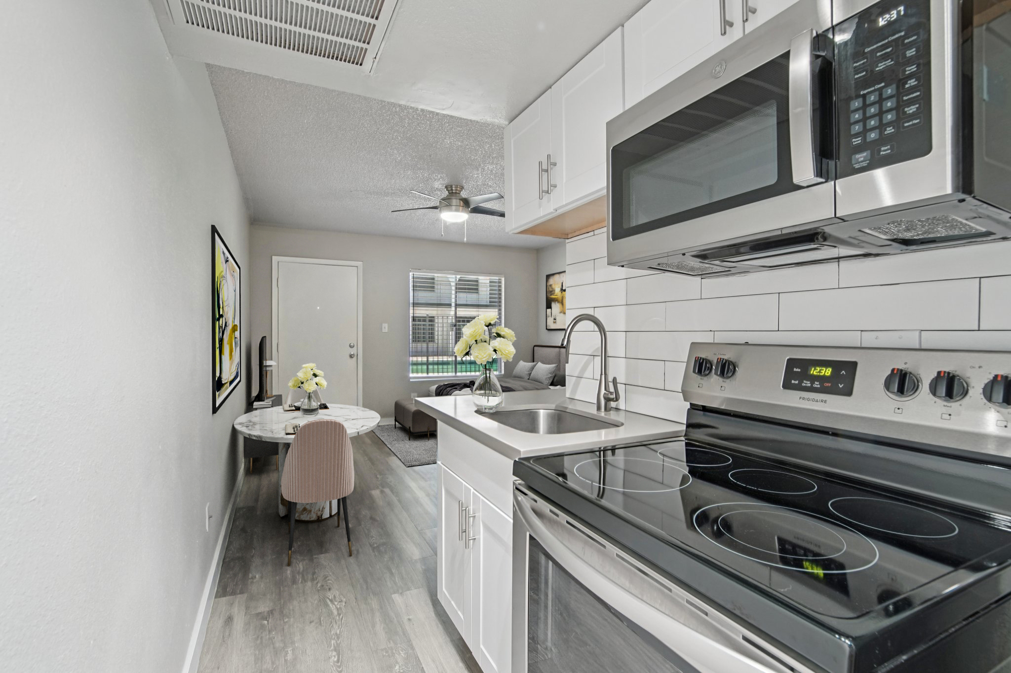 A modern kitchen featuring stainless steel appliances, including a stove and microwave, with white cabinets and a sink. Adjacent to the kitchen is a small dining area with a round table and a chair, and a cozy living room visible in the background. Natural light illuminates the space.
