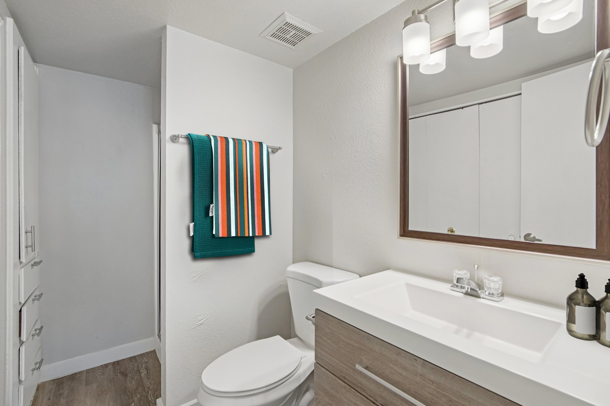 A modern bathroom featuring a white sink with a wooden cabinet below, a toilet, and a large mirror above the sink. A colorful striped towel hangs on a rack, and there are decorative items on the sink. The walls are painted light gray, and the floor has wooden planks.