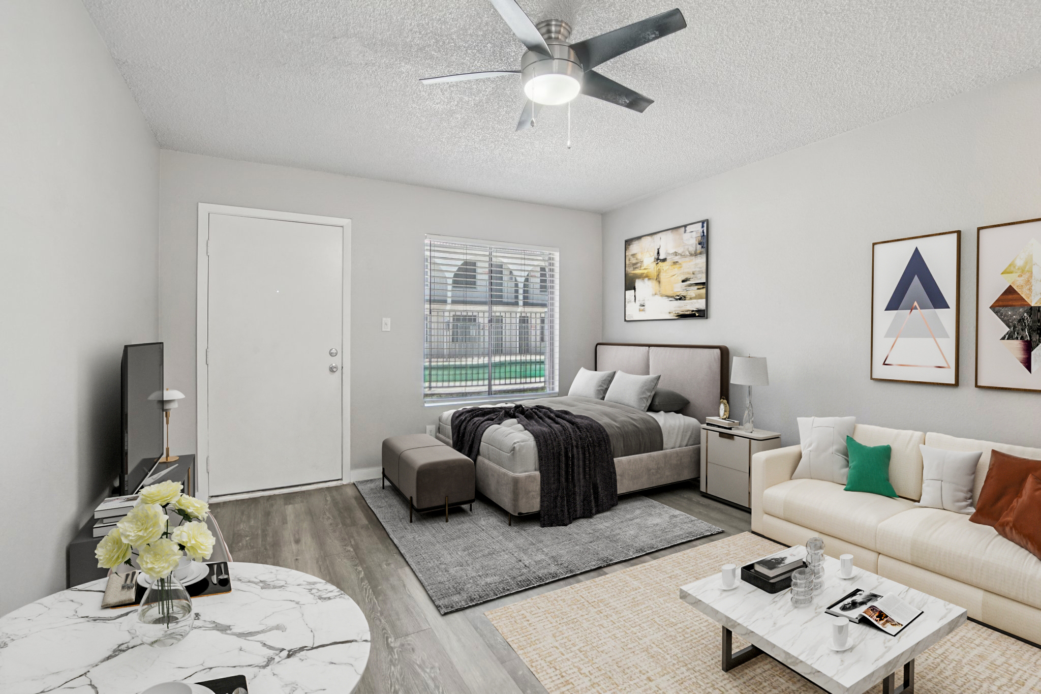 A modern, cozy studio apartment featuring a bed with dark bedding, a stylish sofa, a round marble table with flowers, and a flat-screen TV. The room has light gray walls, a ceiling fan, and a window with an outdoor view. Decor includes abstract artwork on the walls and a soft area rug.