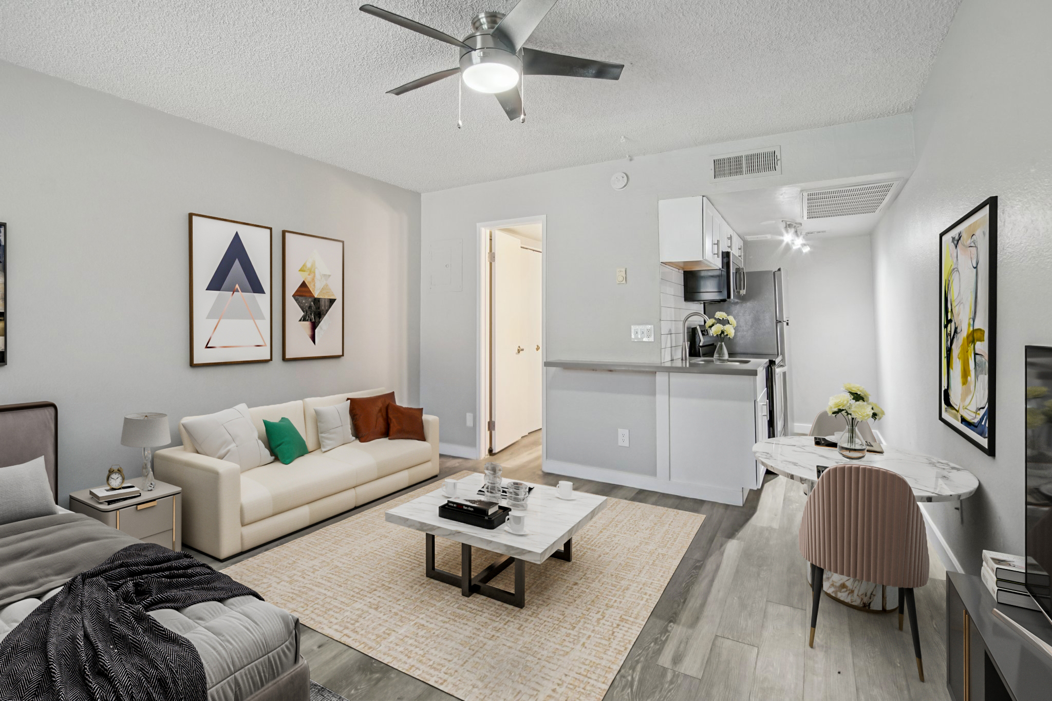 Modern living room featuring a white leather sofa, decorative cushions, and abstract wall art. A light-colored rug covers the floor, with a coffee table at the center. The kitchen is partially visible, showcasing stainless steel appliances. A ceiling fan adds to the contemporary feel, complemented by neutral wall colors.