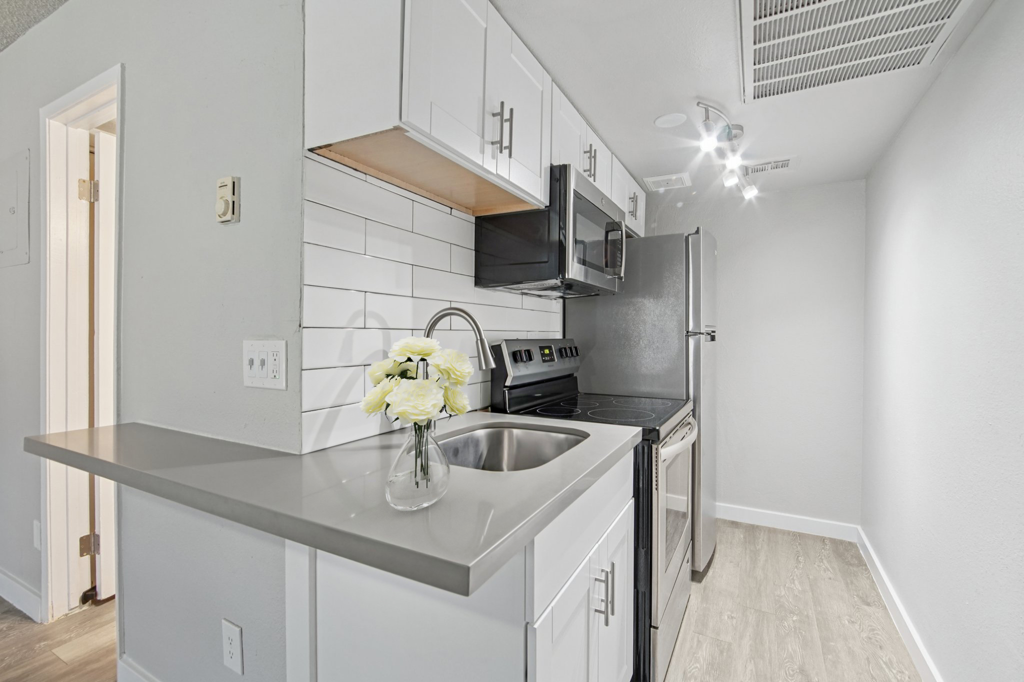 Modern kitchen featuring white cabinetry, a stainless steel microwave and oven, and a silver refrigerator. The counter has a sleek design with a small sink. A vase with white flowers adds a decorative touch. The space has neutral gray walls and bright lighting, creating a clean and inviting atmosphere.
