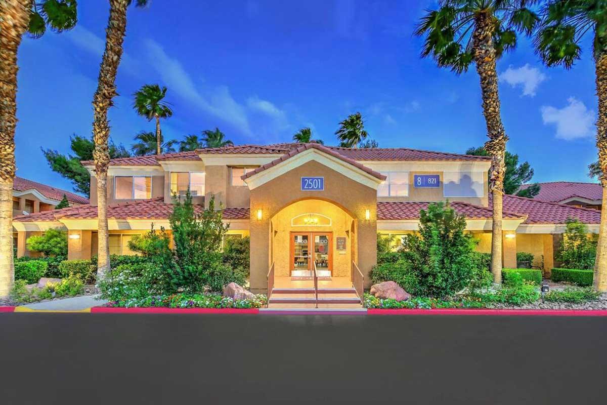 A welcoming entrance of a building with a tan facade, red tiled roof, and palm trees surrounding it. The building features large windows, potted plants, and a well-lit doorway. A sign displays the number "2501" prominently near the entrance. The scene is set during twilight with a clear blue sky.