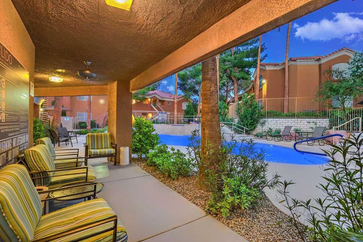 A cozy patio area with striped chairs overlooks a swimming pool surrounded by palm trees and green shrubs. The evening sky reflects a soft light, enhancing the serene atmosphere of the space. The pool area is well-maintained, offering a relaxing retreat in a residential setting.