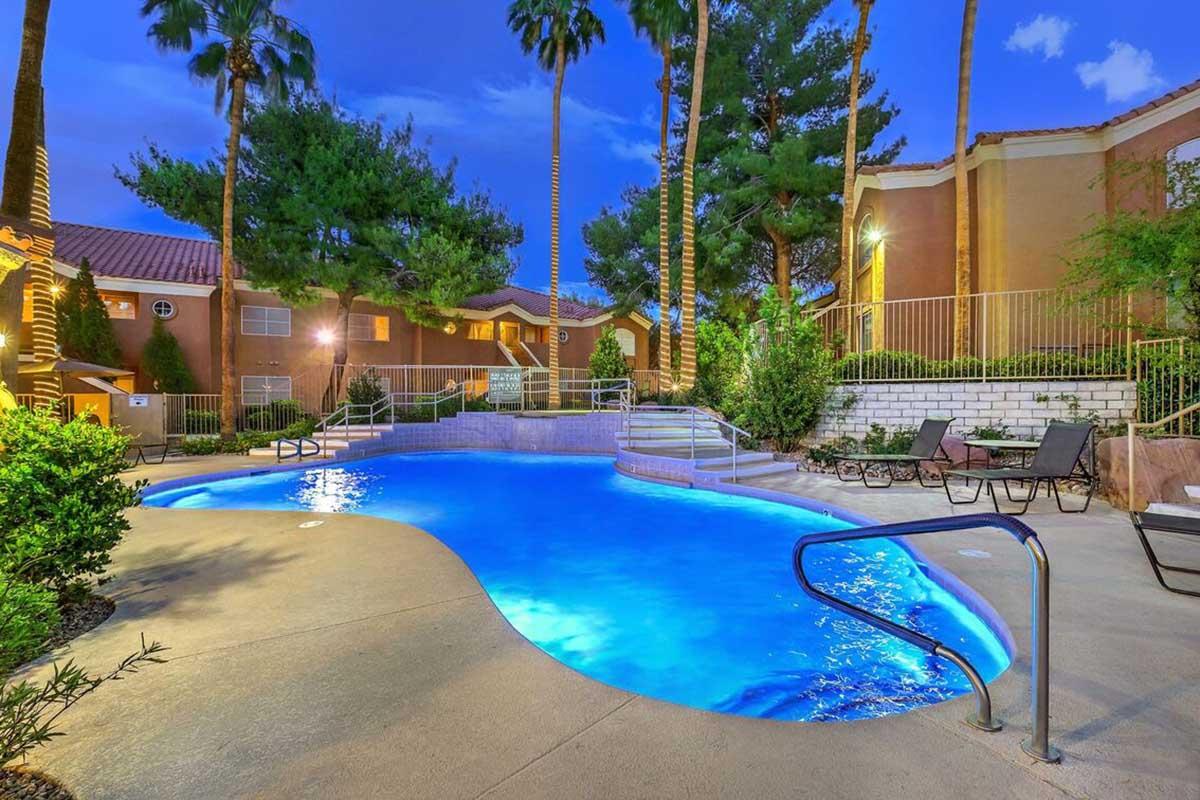 A beautifully lit outdoor swimming pool surrounded by palm trees and lush greenery, with lounge chairs nearby. The scene is set during twilight, showcasing the calming blue water and the warm glow of lights illuminating the area. A tranquil environment perfect for relaxation.