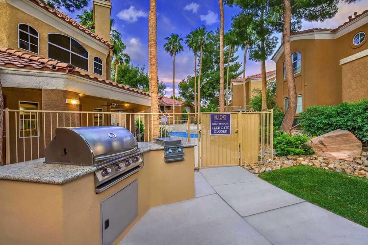 A well-maintained outdoor area featuring a stainless steel grill next to a gated pool entrance, surrounded by palm trees and colorful landscaping, with two adjacent buildings visible in the background under a blue sky.