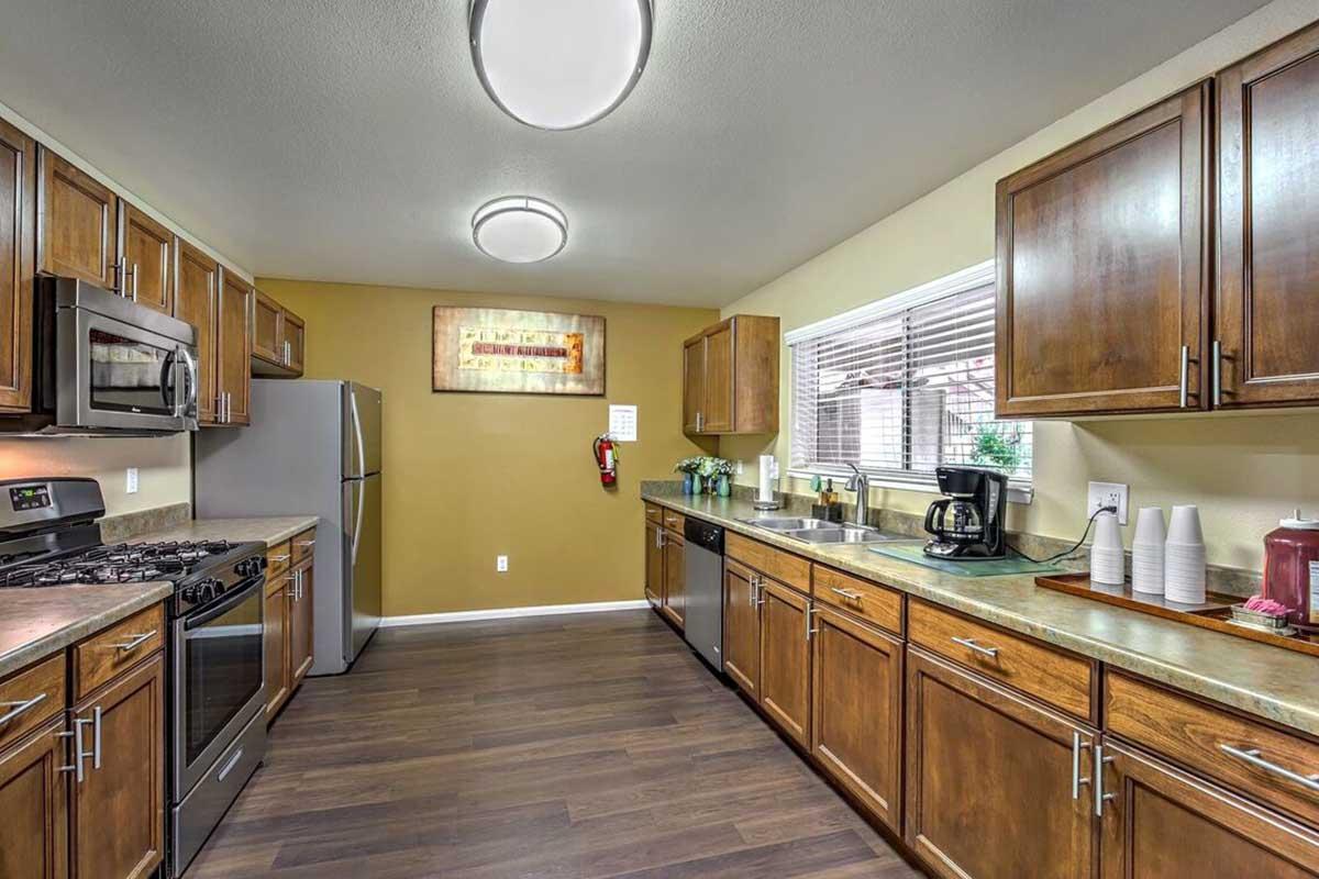 A well-lit kitchen featuring wooden cabinets, stainless steel appliances including a refrigerator and oven, and a countertop with coffee maker and cups. The walls are painted in warm tones, and there's a window allowing natural light to enter, enhancing the inviting atmosphere.