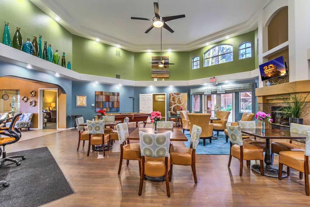 A spacious interior of a modern lounge or community area featuring multiple tables with chairs, decorative plants, and colorful vases on display. Large windows allow natural light to brighten the space, which includes seating areas and a pool table, creating a welcoming social atmosphere.