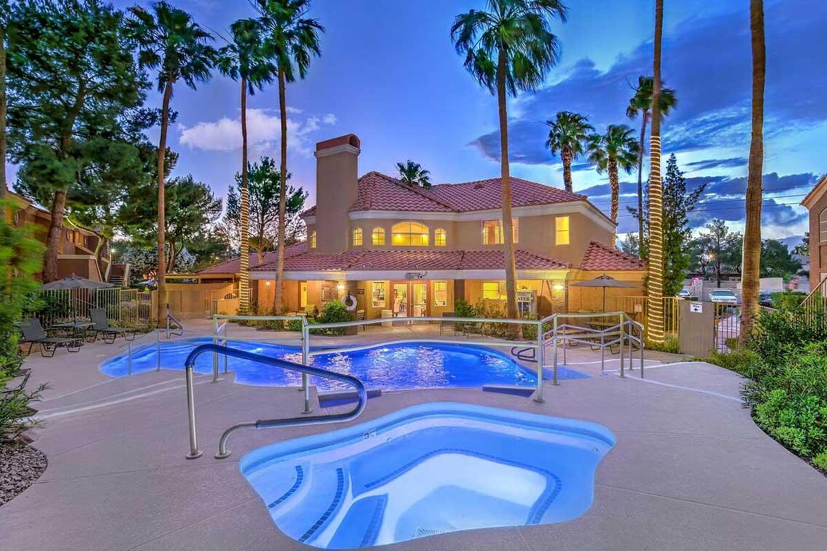 A beautifully landscaped pool area featuring a large swimming pool and a hot tub, surrounded by palm trees. The sun is setting, casting a warm glow over the scene. In the background, a spacious building with a red tile roof and large windows is visible, enhancing the inviting atmosphere of the setting.